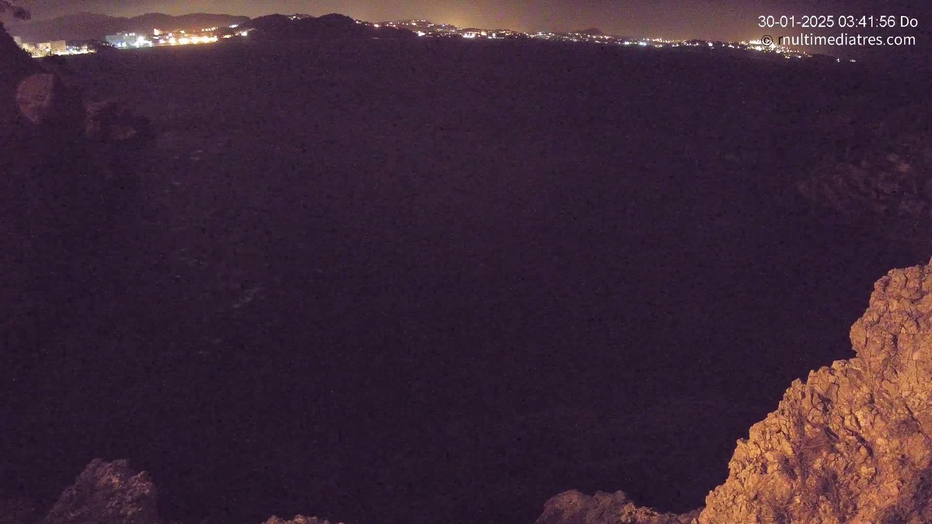 A nighttime view from a rocky outcrop overlooking a dark body of water and a distant city skyline illuminated by lights.