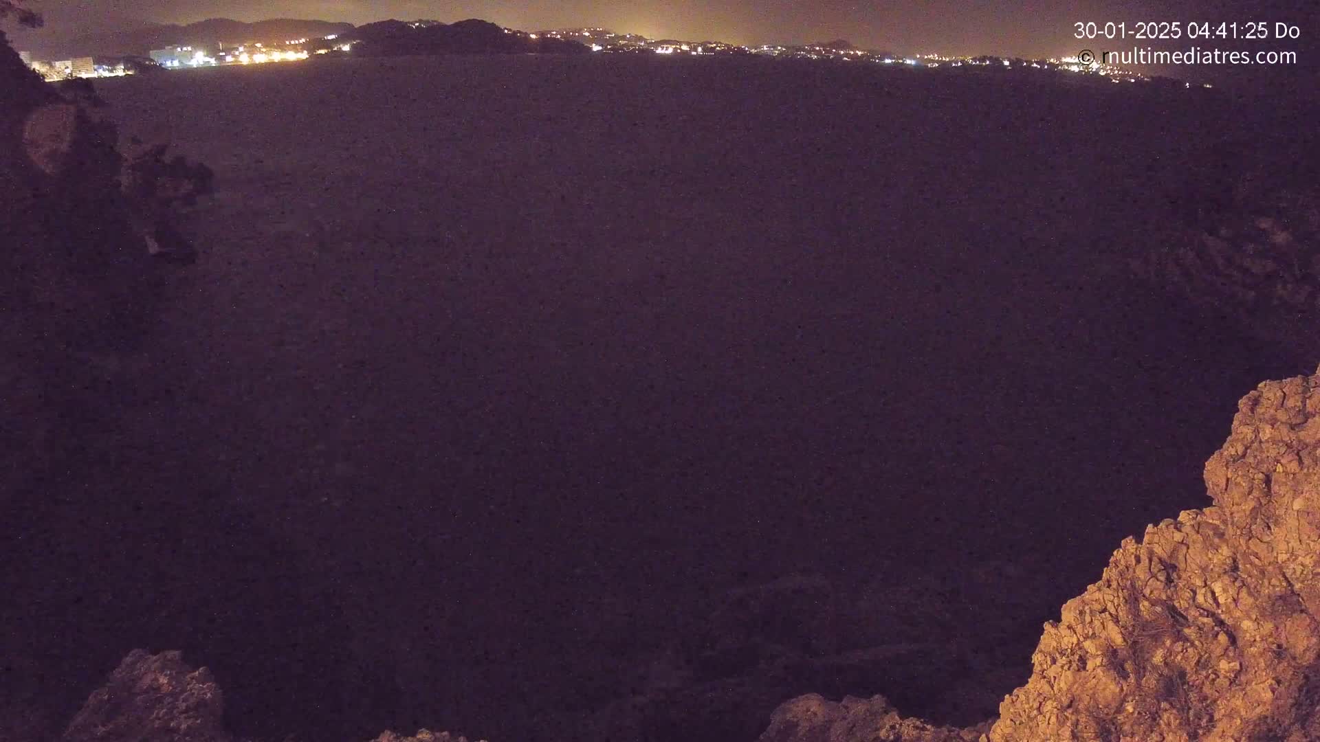 A nighttime view of a dark body of water with a distant city skyline illuminated, partially framed by rocky terrain.