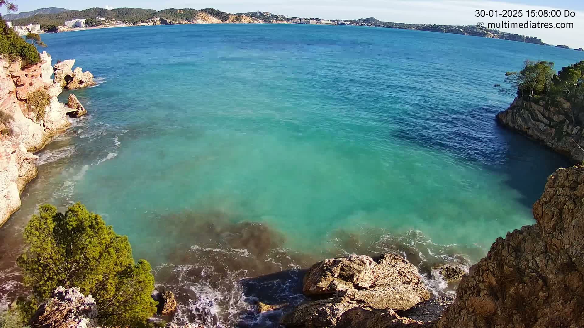 Bright turquoise water laps against rocky cliffs on a sunny day, with a distant coastline visible in the background.
