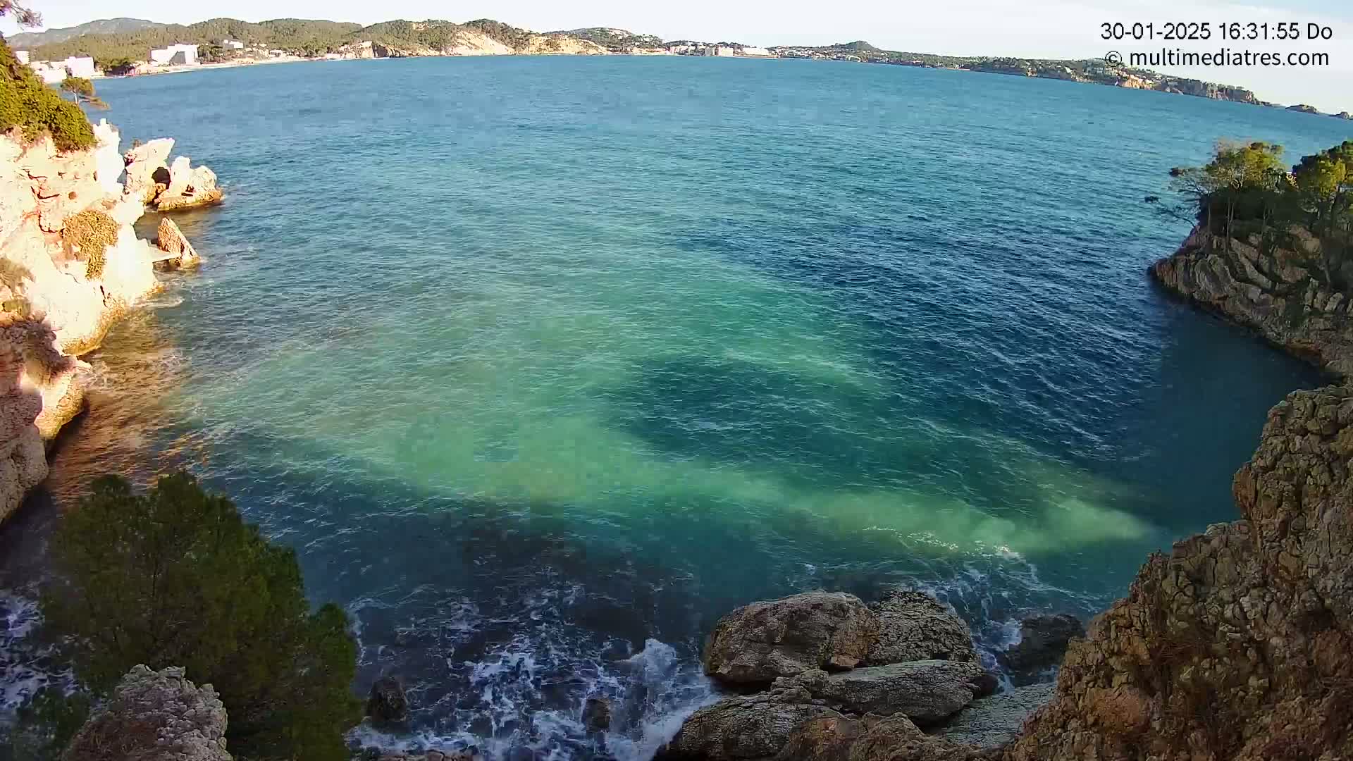 Rocky cliffs enclose a cove of clear, turquoise water lapping gently against the rocks under a sunny sky.