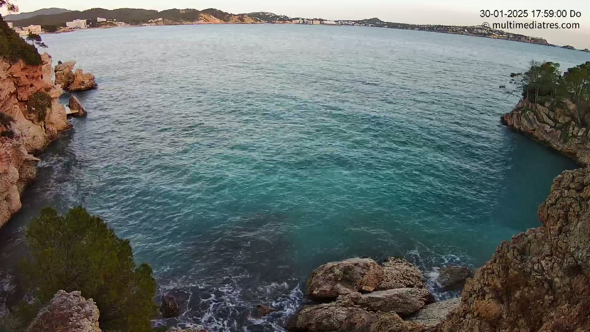A tranquil cove of teal water is nestled between rocky cliffs, with a distant coastline visible under a calm, twilight sky.