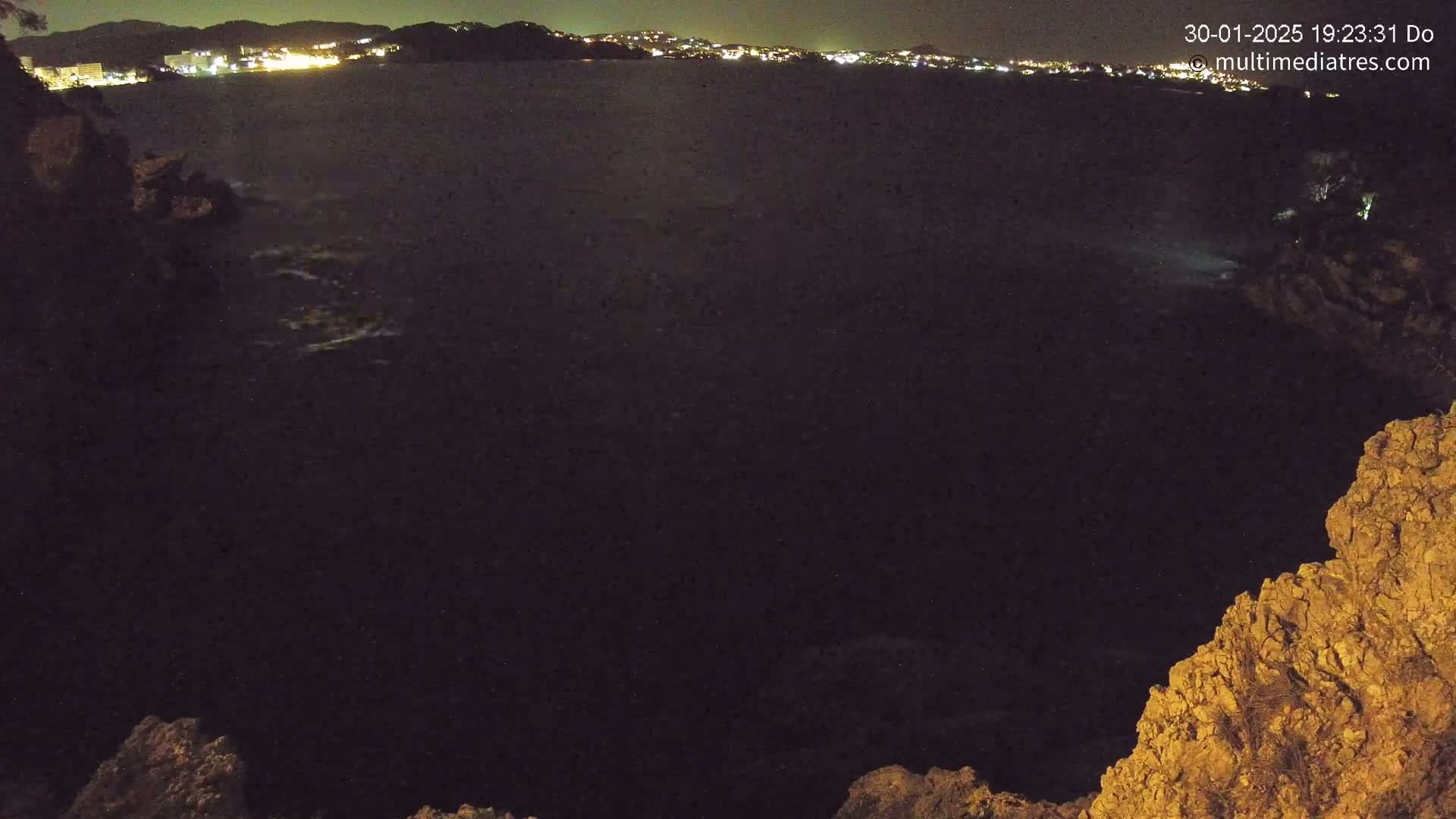 A dark, nighttime view of a rocky coastline with a body of water and a distant, illuminated town visible in the background.