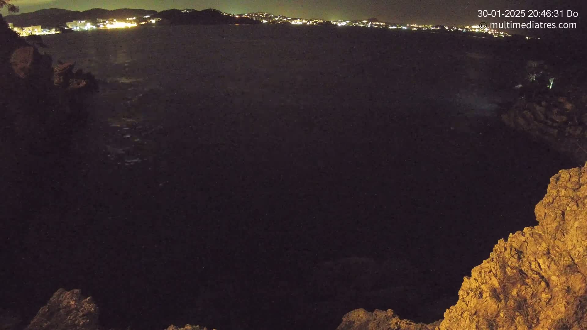 A nighttime view from a rocky cliff overlooking a dark body of water, with a distant city illuminated on the horizon.