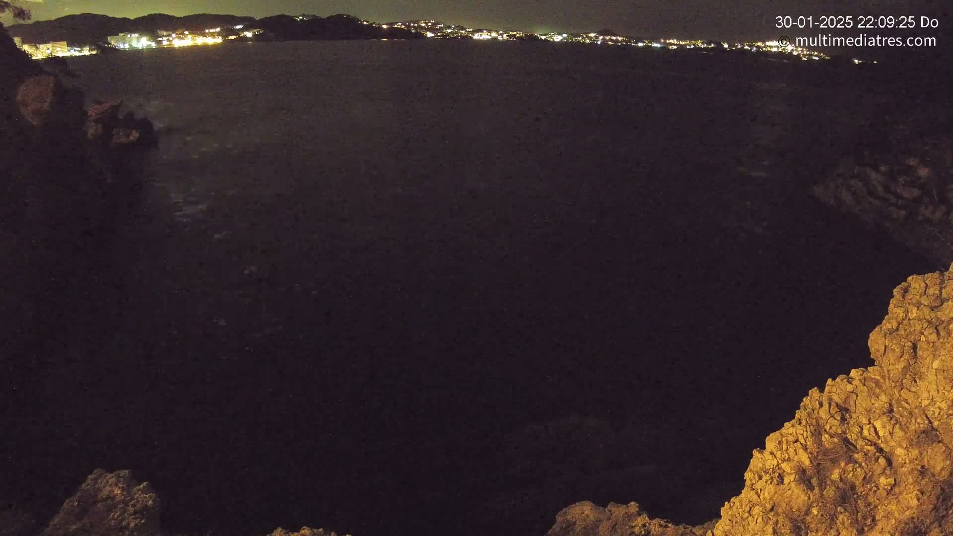 A nighttime view of dark water, possibly an ocean or sea, with a distant city skyline illuminated by lights, framed by rocky terrain.