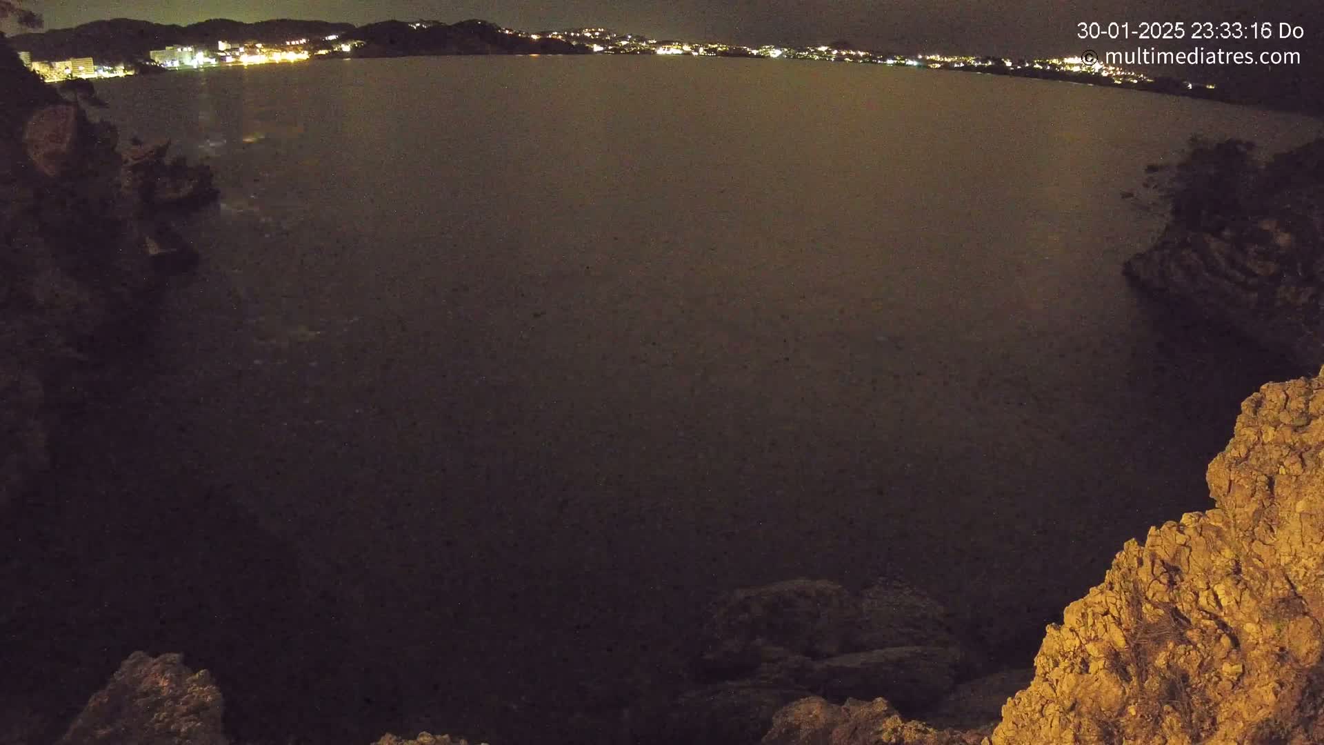 A nighttime, dark-colored body of water is seen from a high vantage point, with rocky terrain in the foreground and a distant, faintly lit shoreline in the background.