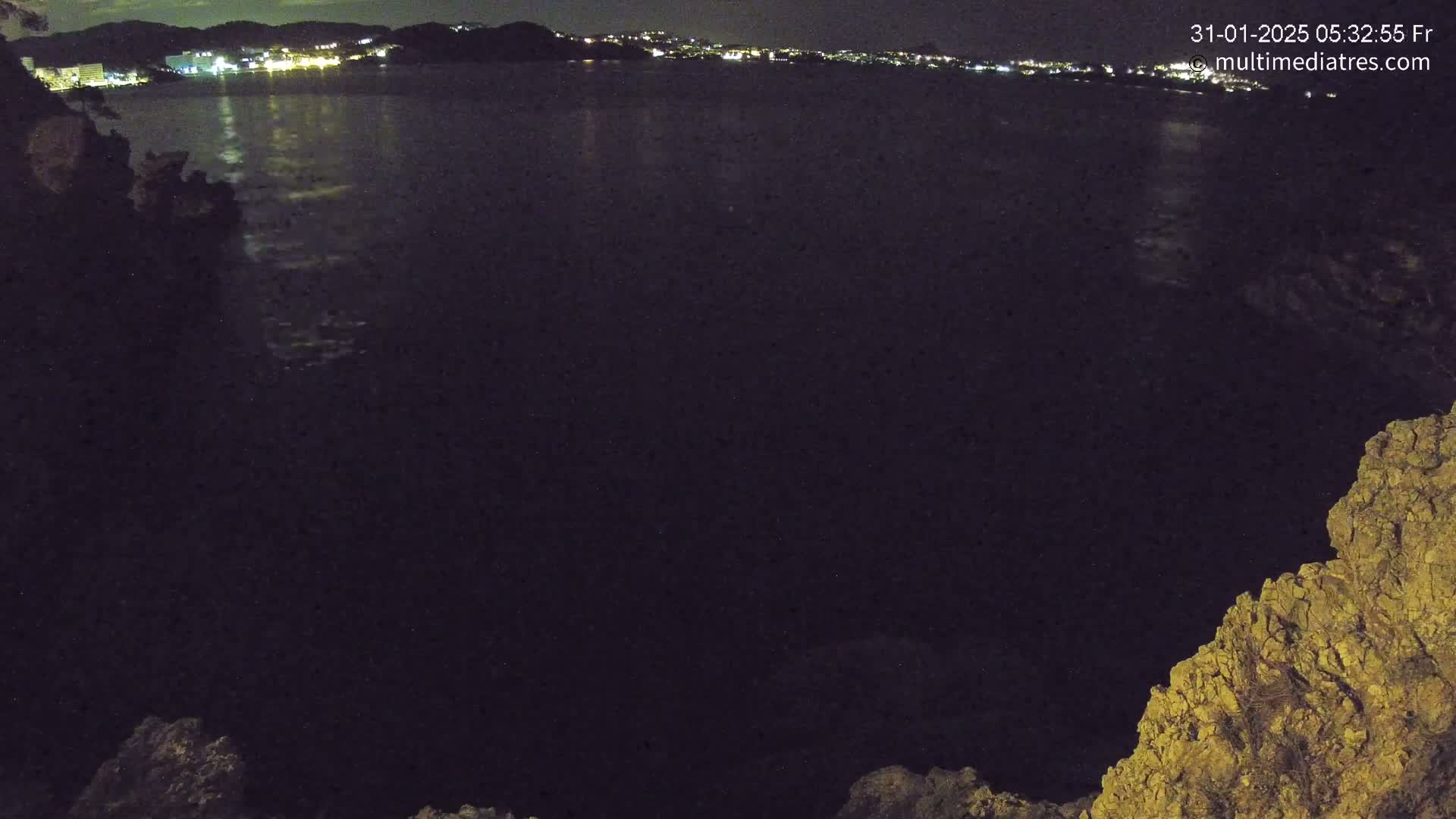 A nighttime view of a dark body of water reflecting the lights of a distant town, seen from a rocky, elevated vantage point.