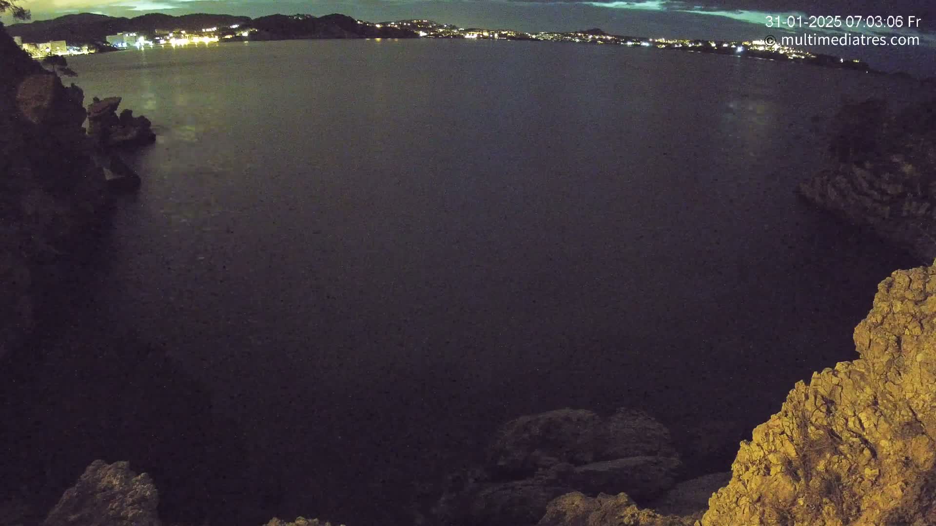 A nighttime, high-angle view shows a dark body of water with a distant, faintly lit shoreline, partially framed by rocky terrain.