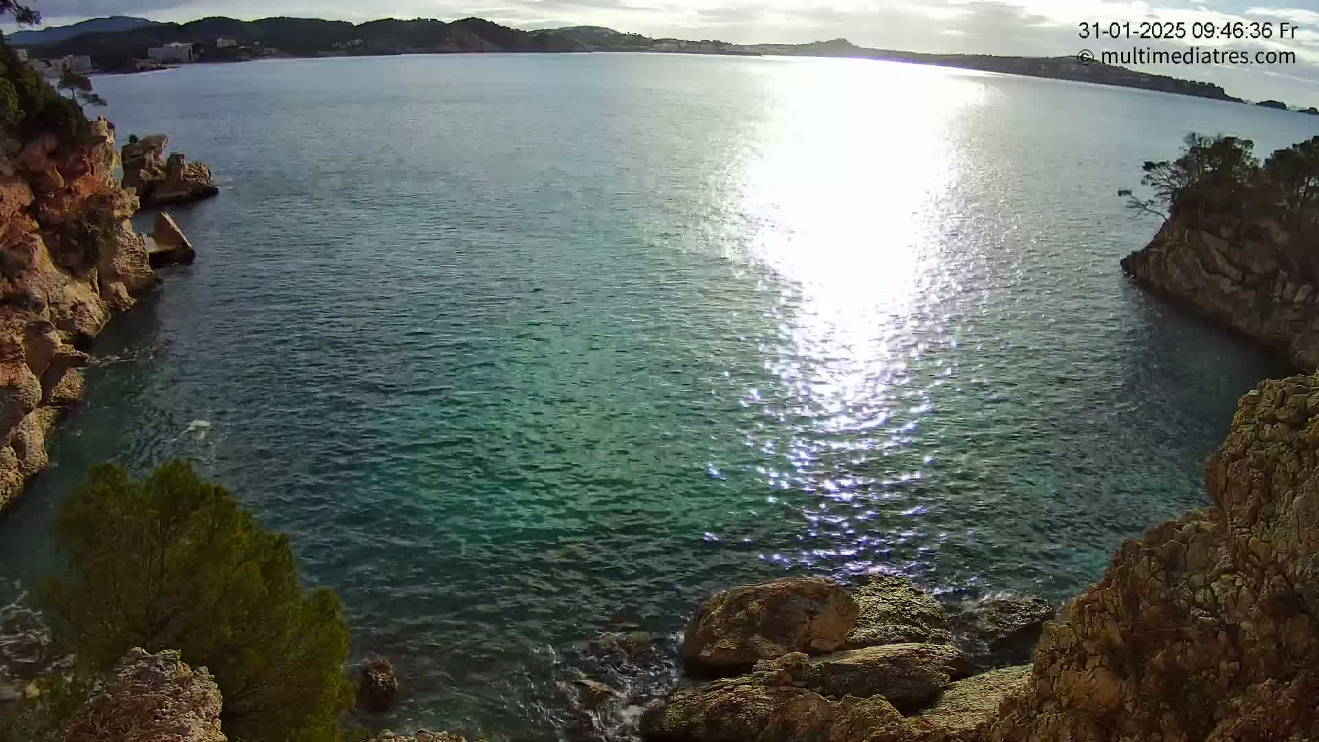 Sunlight glistens on the calm, teal-colored water of a cove nestled between rocky cliffs on a partly sunny day.