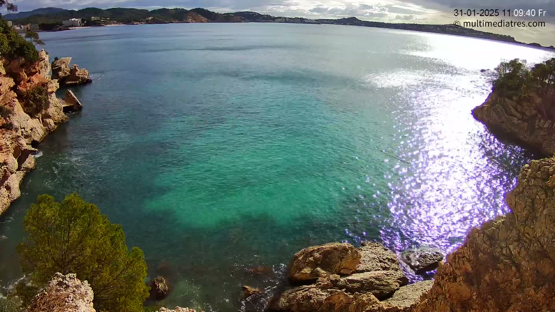 Clear, teal water laps against rocky, tan cliffs under a partly cloudy sky.