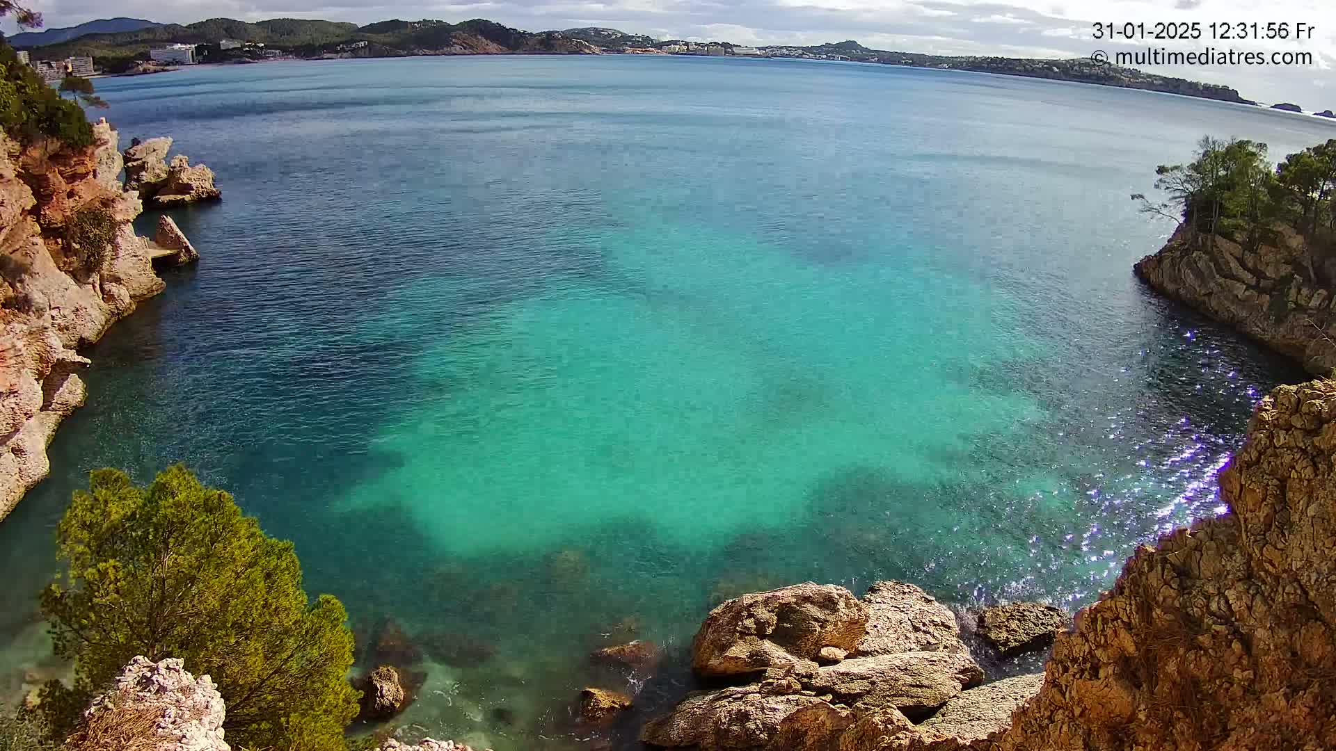 A cove of clear, turquoise water is nestled between rocky, tan-colored cliffs under a partly sunny sky.
