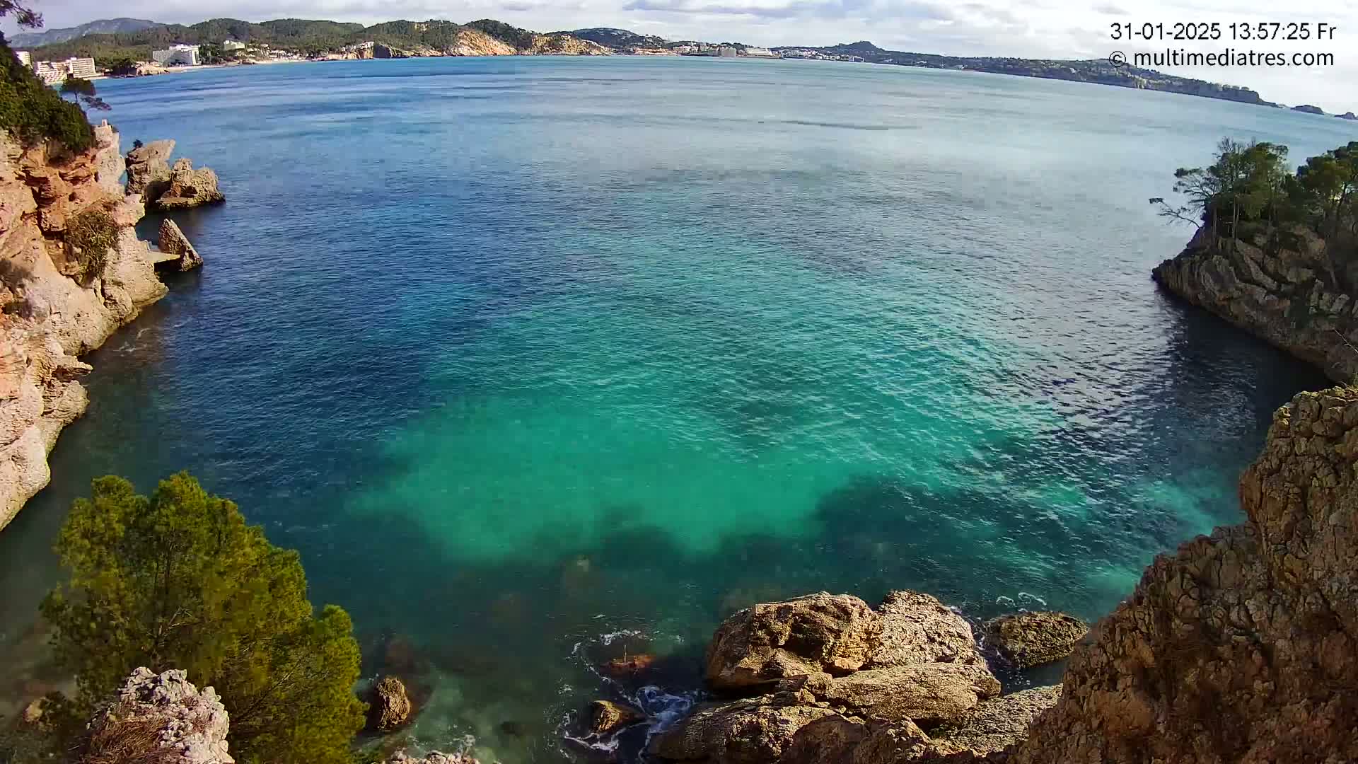 A cove of clear, teal water is nestled between rocky, tan cliffs under a partly cloudy sky.