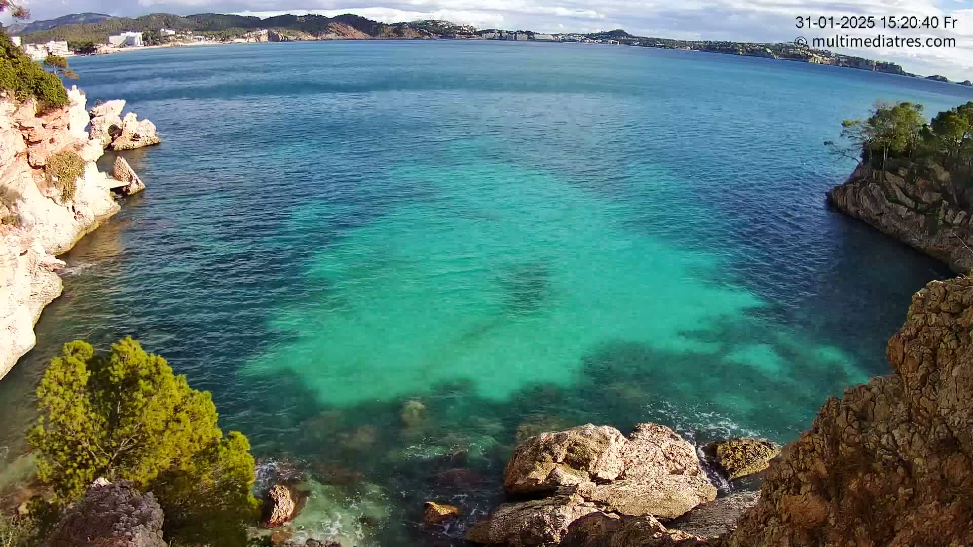 Bright turquoise water fills a cove between rocky cliffs, with a distant town visible across the calm bay under a partly cloudy sky.