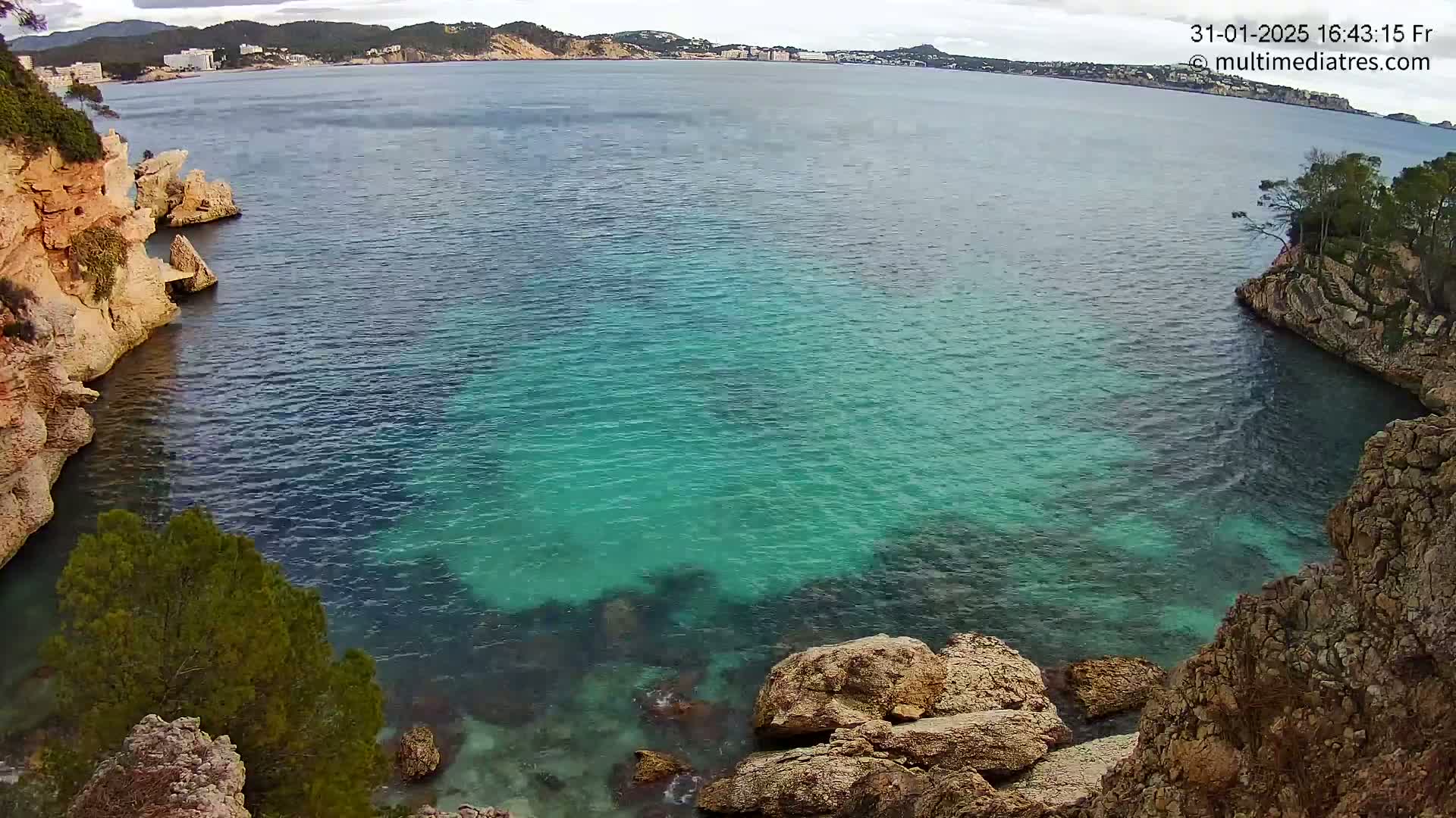A cove of teal-colored water is nestled between rocky, tan-colored cliffs on a partly cloudy day, with a town visible in the distance.