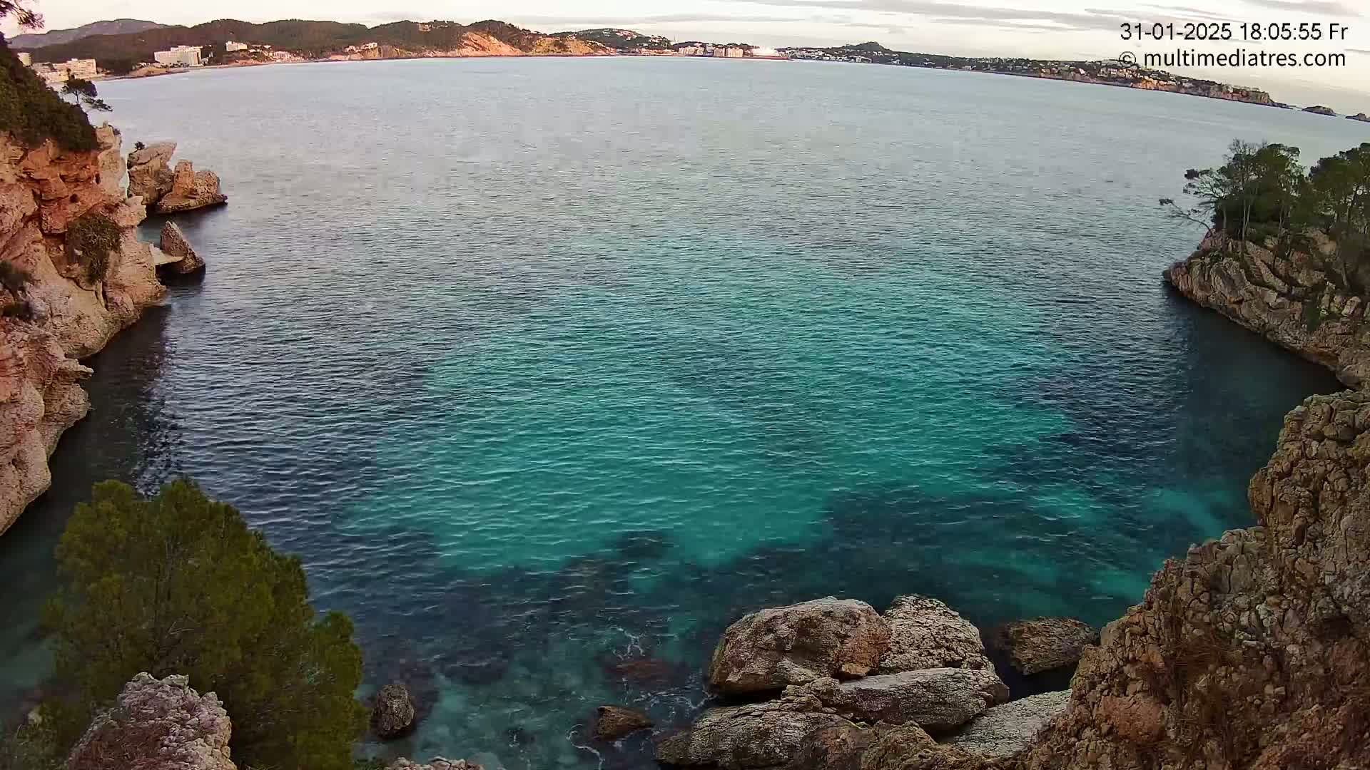 A tranquil cove of teal water is nestled between rocky cliffs, with a distant coastline visible under a partly cloudy sky.