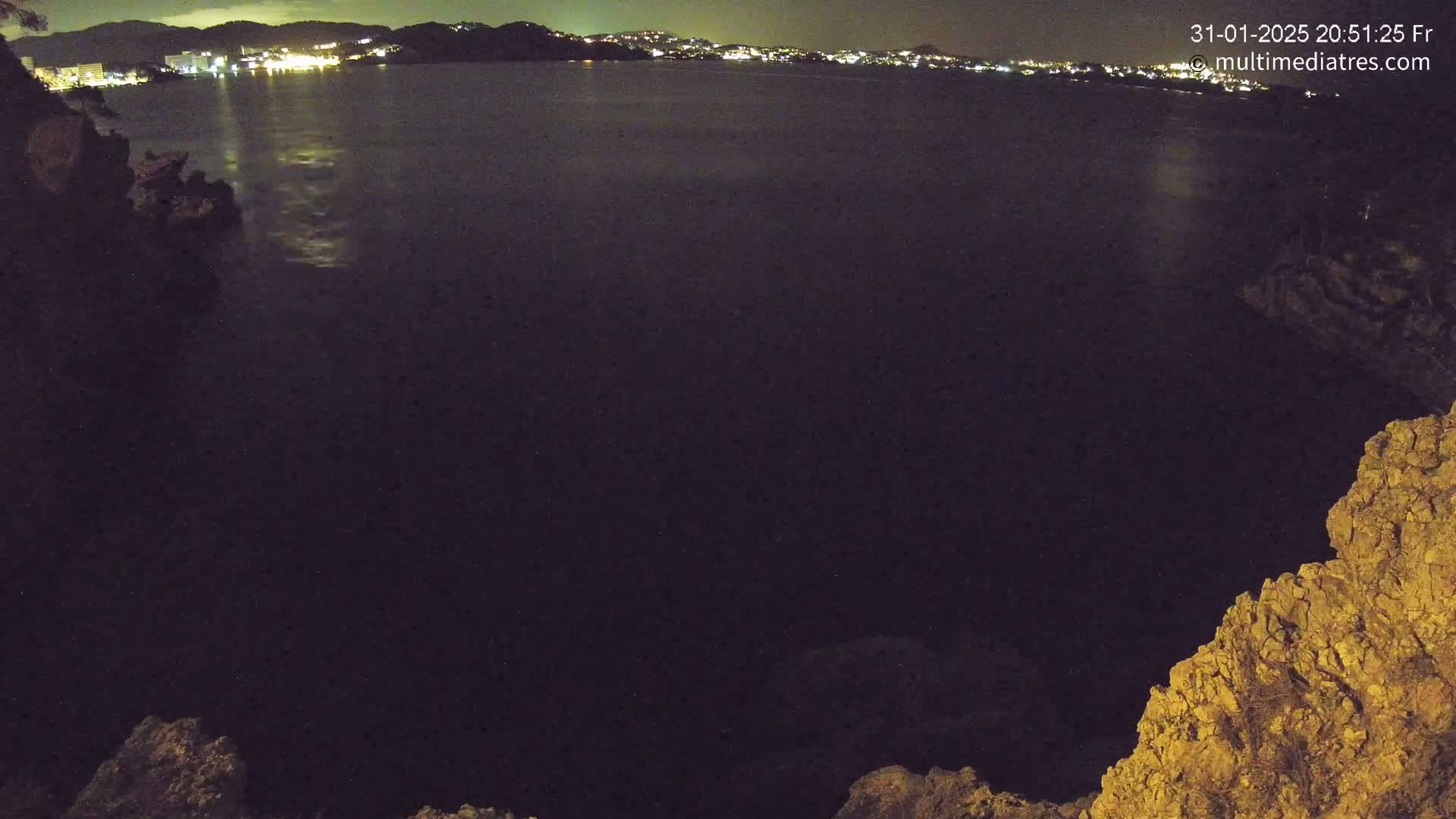 A nighttime view of a dark body of water with a distant, illuminated cityscape and rocky foreground under a clear sky.