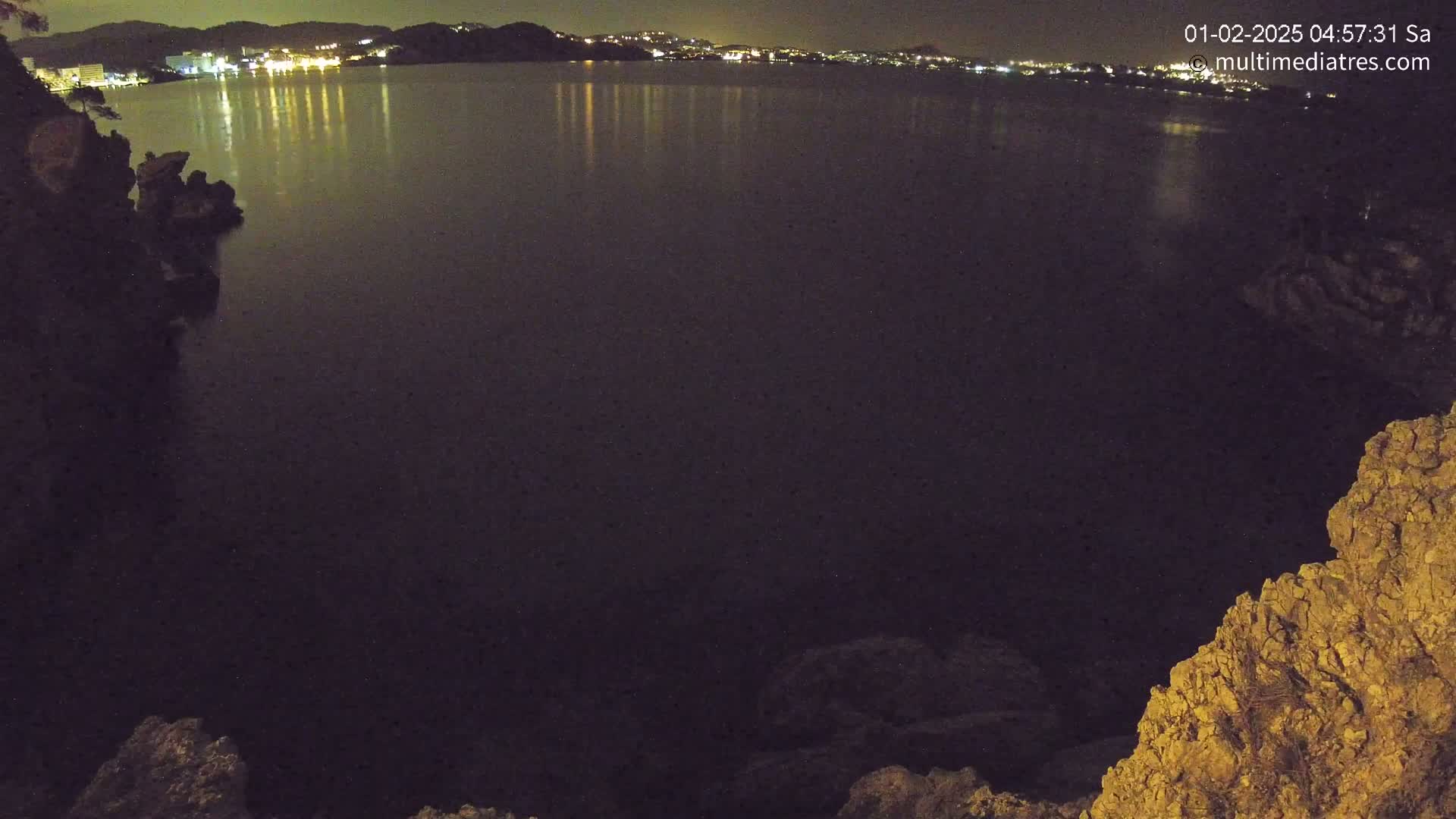 A nighttime view of a dark body of water reflecting the lights of a distant town, seen from a rocky outcrop.