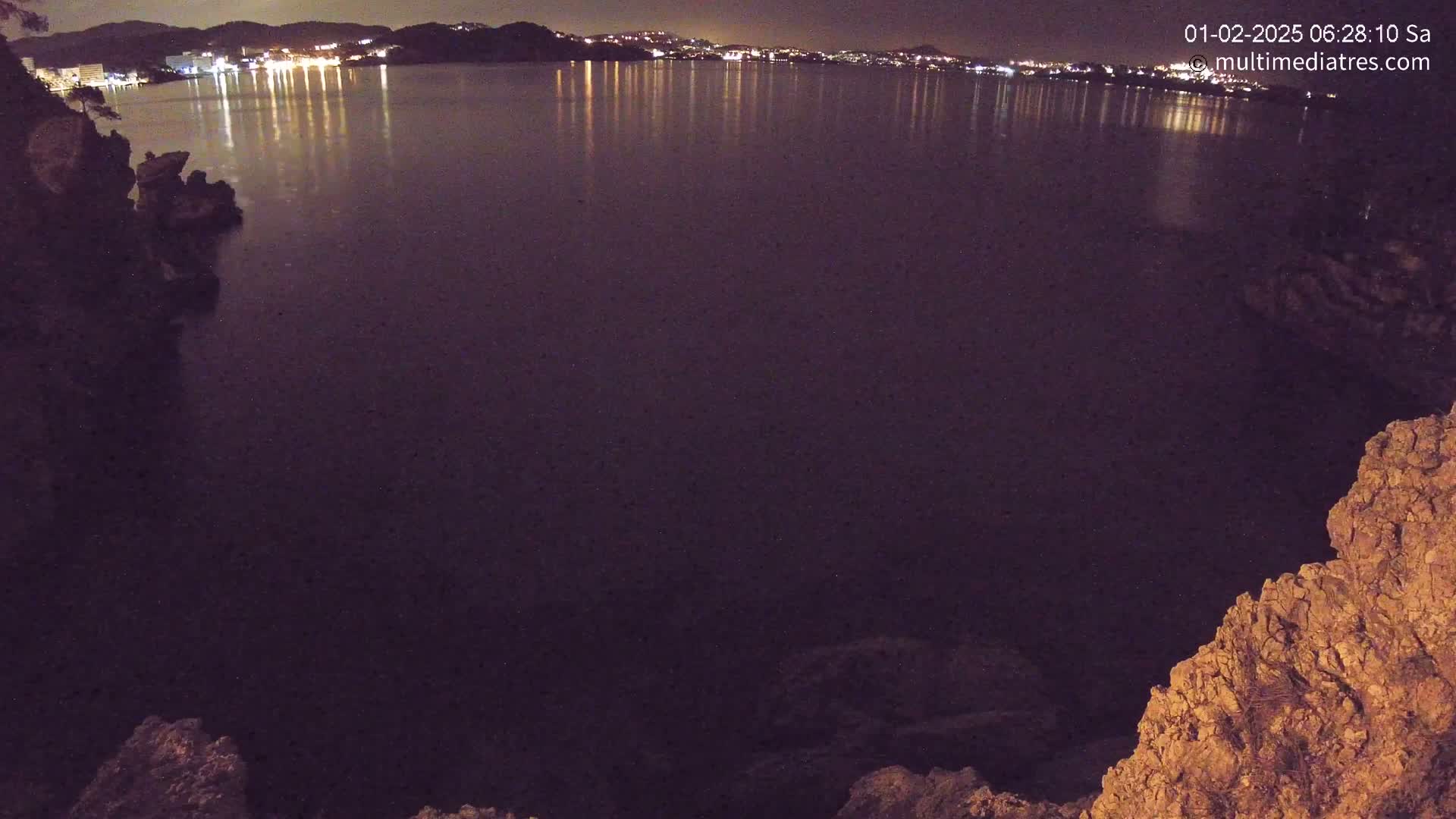 A nighttime view of a dark body of water reflecting the lights of a town across the bay, seen from a rocky cliff.