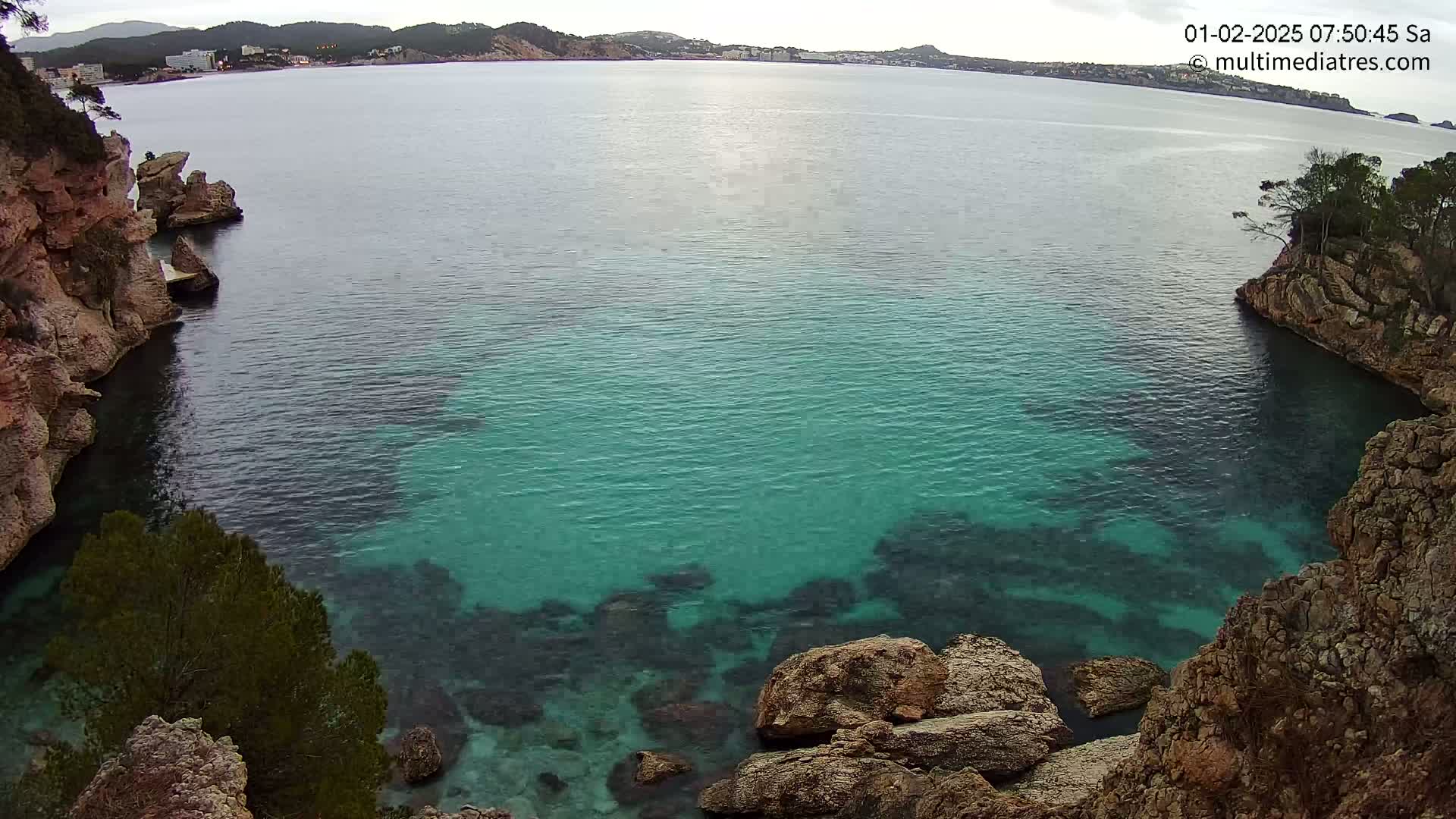 A secluded cove with clear, teal water is nestled between rocky cliffs under an overcast sky.