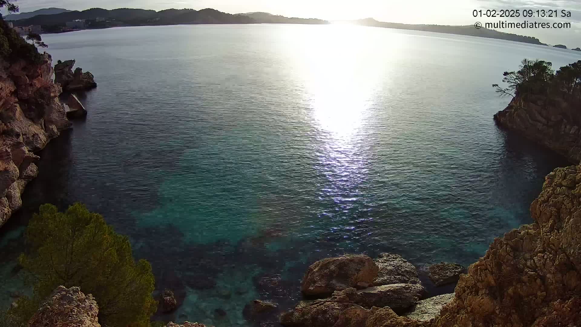 A bright, sunny day overlooks a calm, clear teal ocean cove nestled between rocky, cliff-like shores.