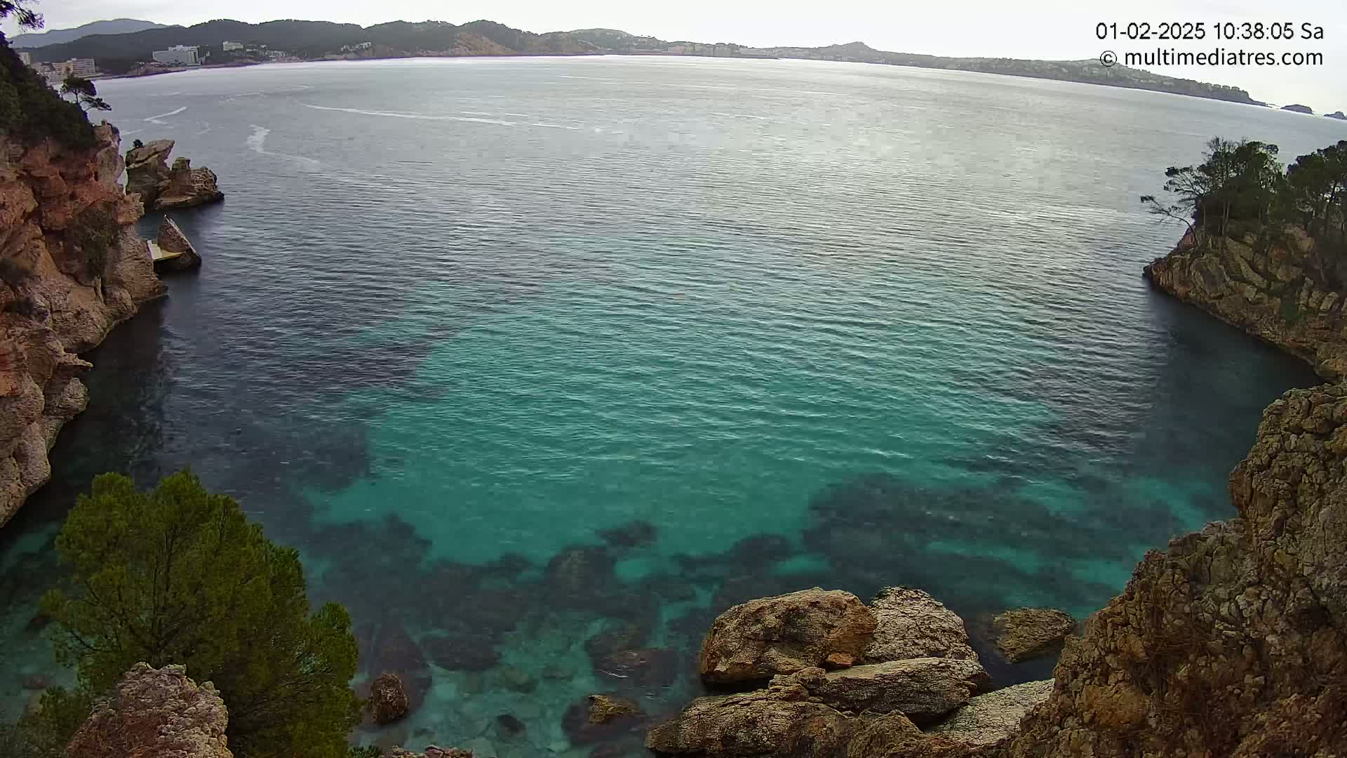 Clear, teal water fills a cove between rocky, tree-covered cliffs on a partly cloudy day.
