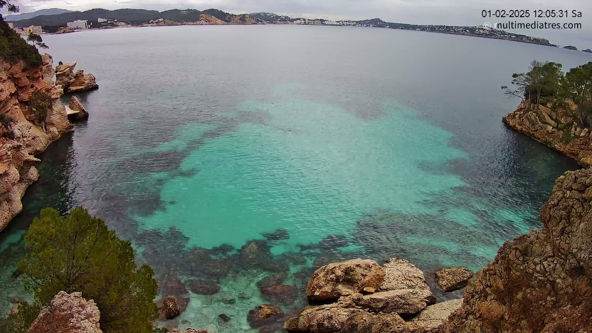 A cove with clear teal water is nestled between rocky, tan-colored cliffs under an overcast sky.