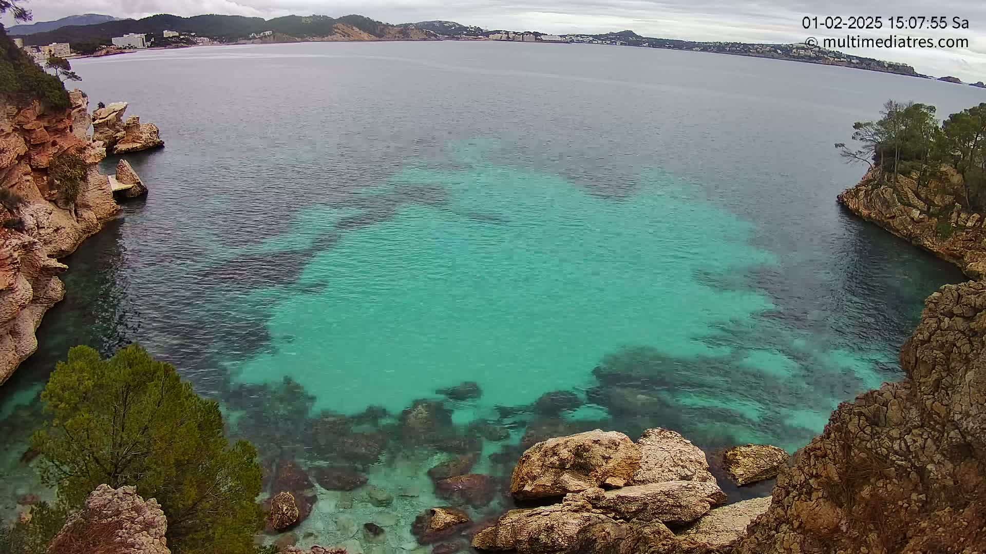 Clear, teal water fills a cove between rocky, tan cliffs under an overcast sky.