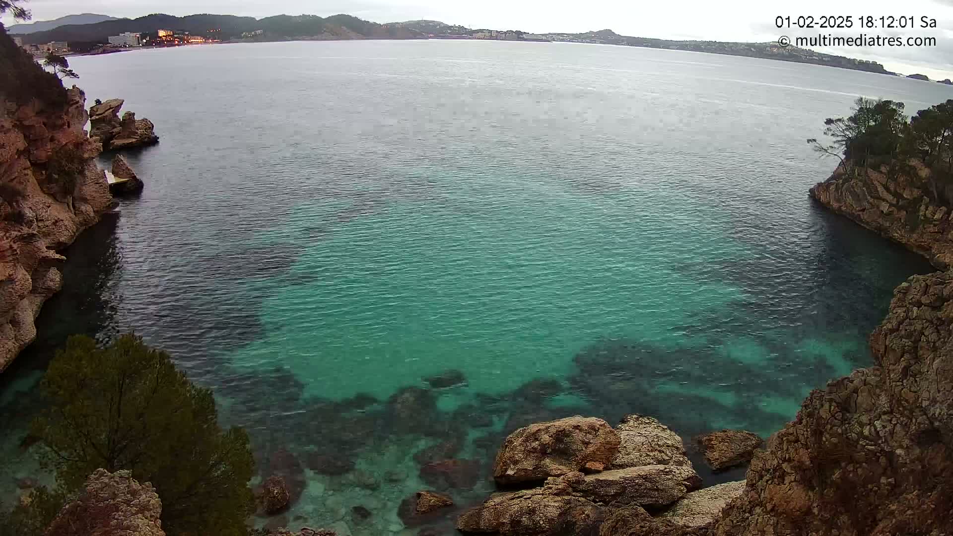 A cove of clear, teal water is nestled between rocky, vegetated cliffs under an overcast sky.