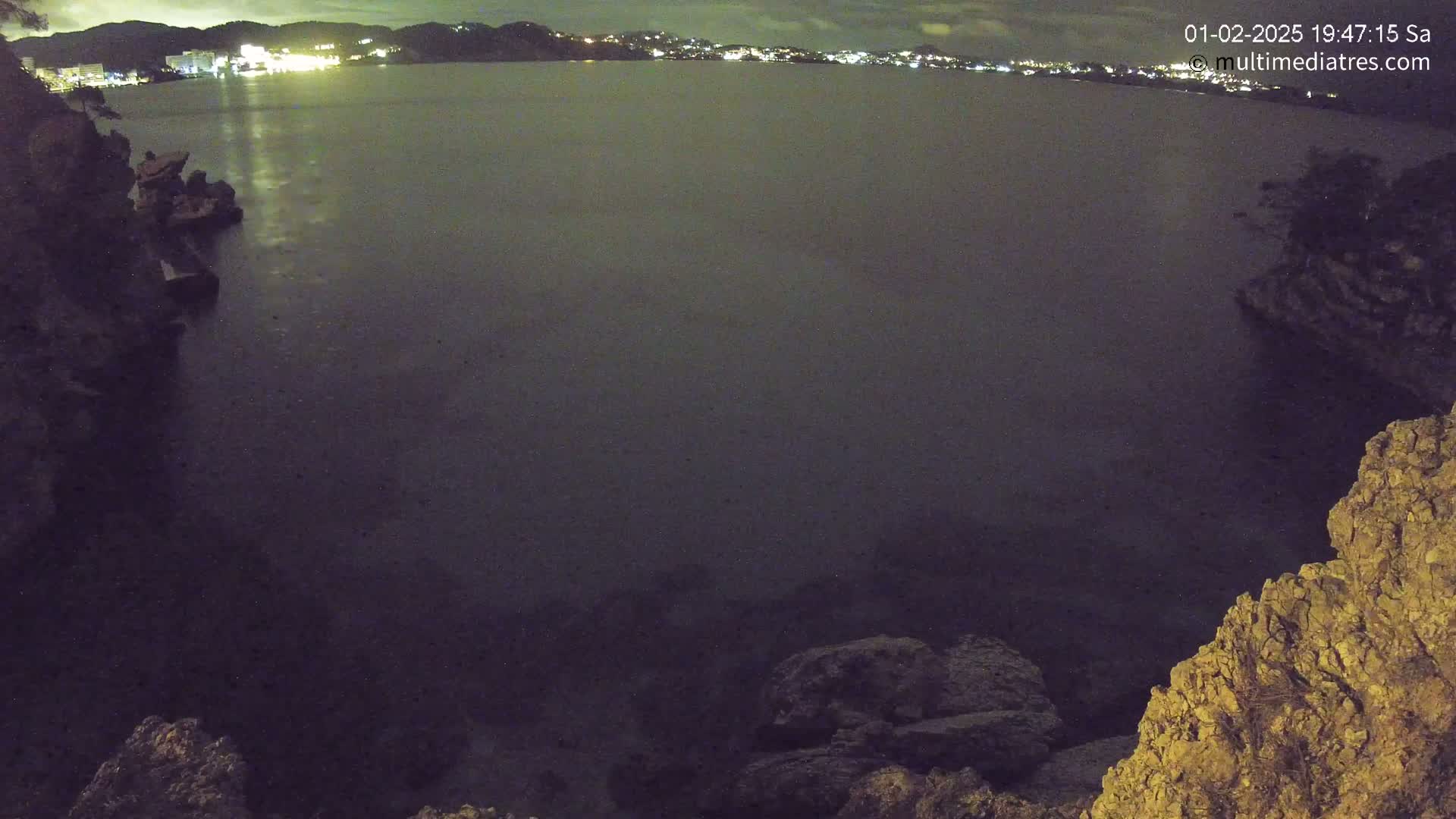 A nighttime view of a dark body of water, with a distant, illuminated cityscape visible on the horizon, framed by rocky, dark land formations.