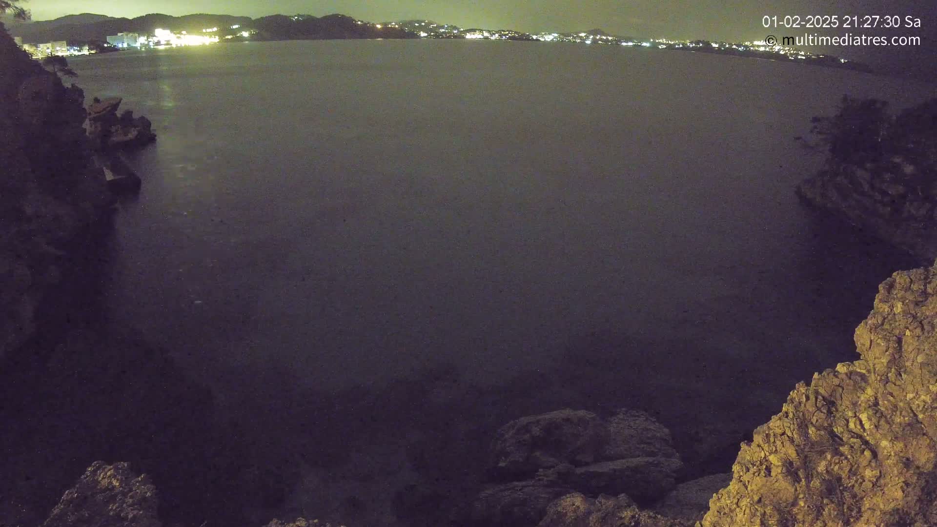 A nighttime view of dark water, a rocky shoreline, and distant lights from a town across the bay.