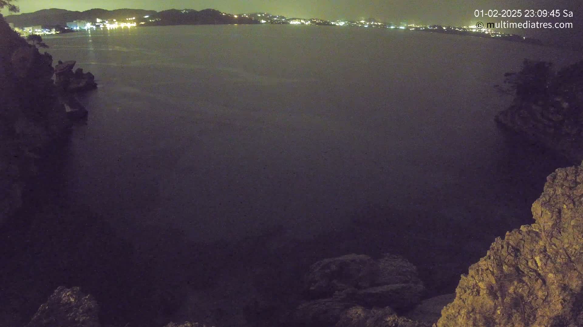 A nighttime view of dark water, illuminated by distant lights on a shoreline, seen from between two rocky outcrops.