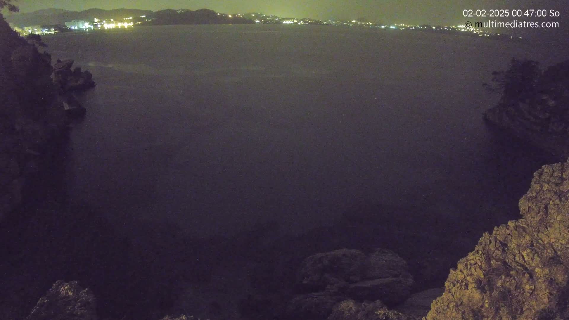 A nighttime view of dark, rocky shoreline with a calm body of water extending to a distant, faintly lit cityscape.