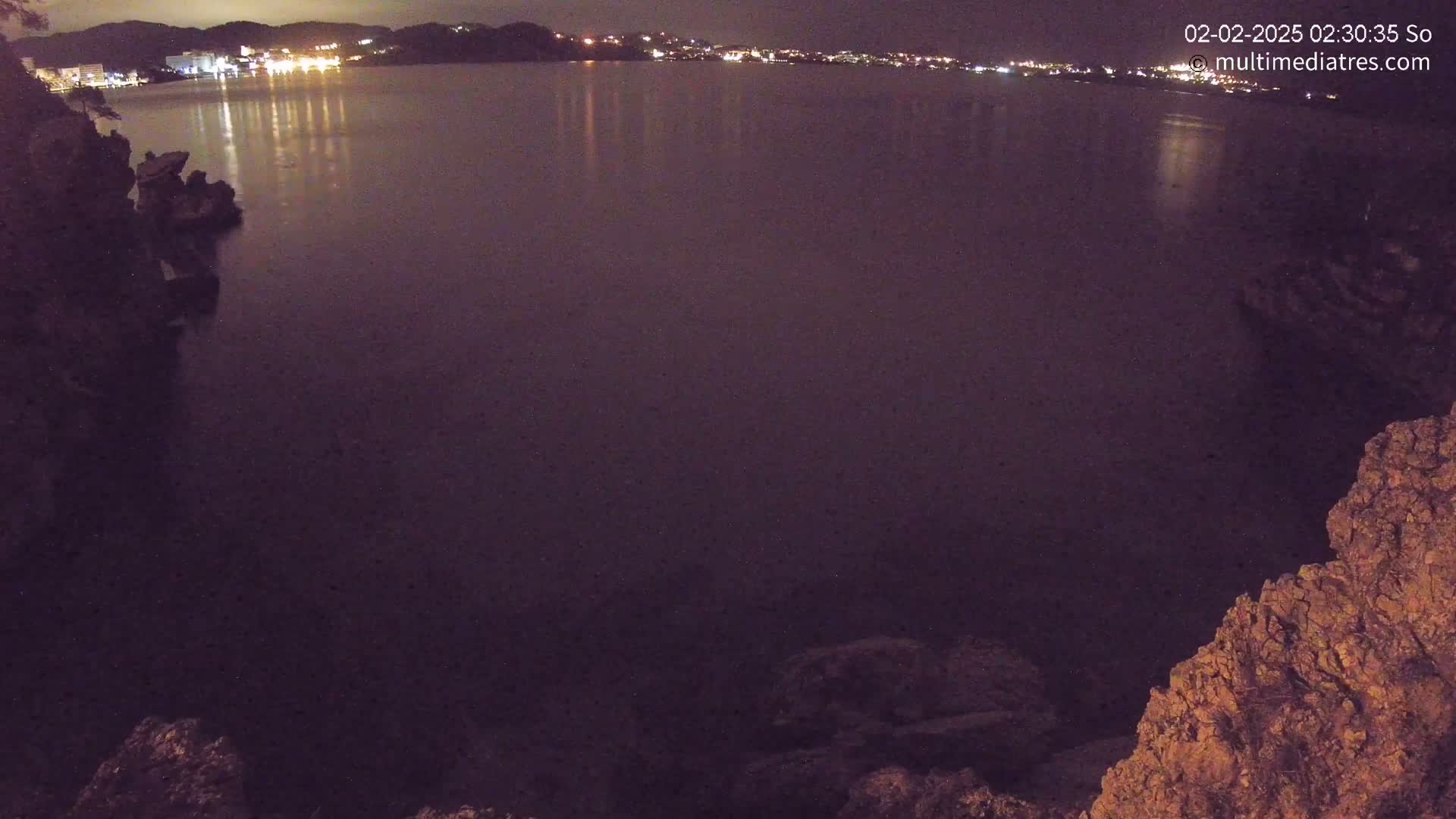 A nighttime view of a calm body of water reflecting city lights across from a rocky shoreline.