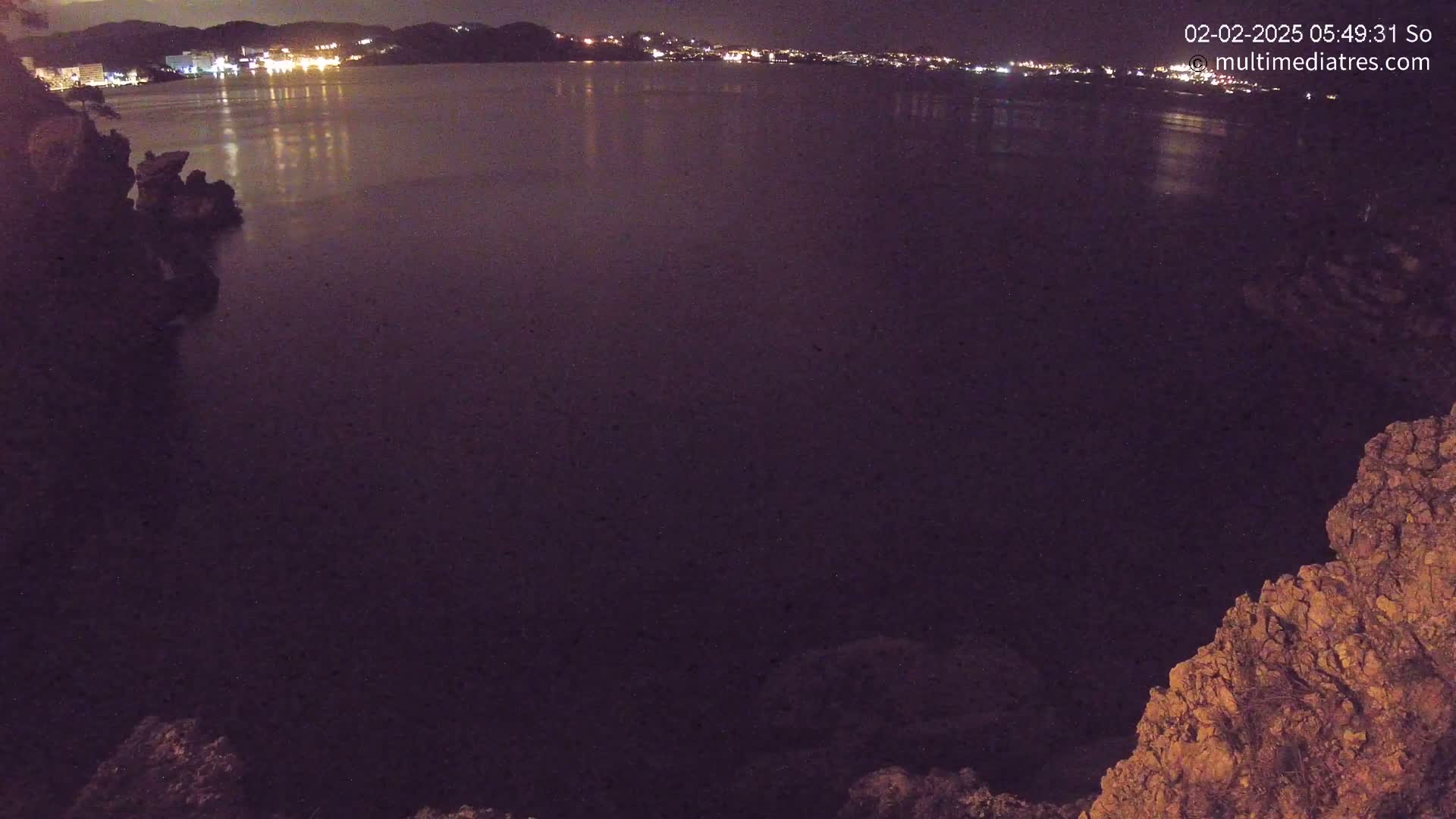A nighttime view of a dark body of water reflecting lights from a distant town, seen from a rocky vantage point.