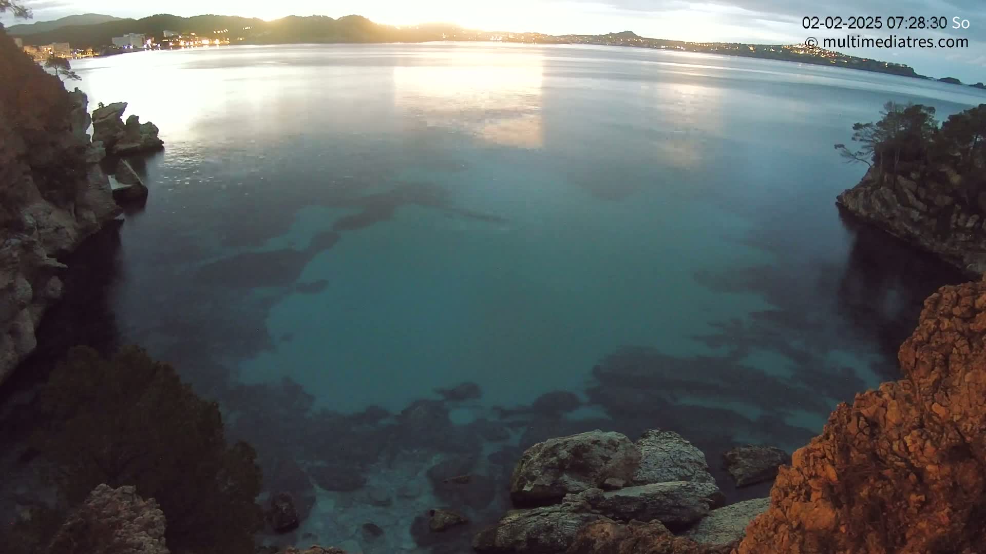 A calm, teal-colored bay is surrounded by rocky cliffs at sunrise, with a town visible in the distance.