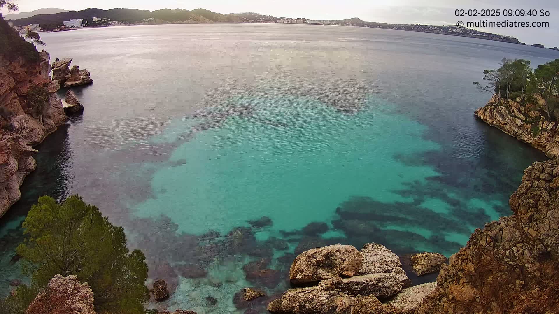 A cove with clear, turquoise water is nestled between rocky, tan cliffs under an overcast sky.