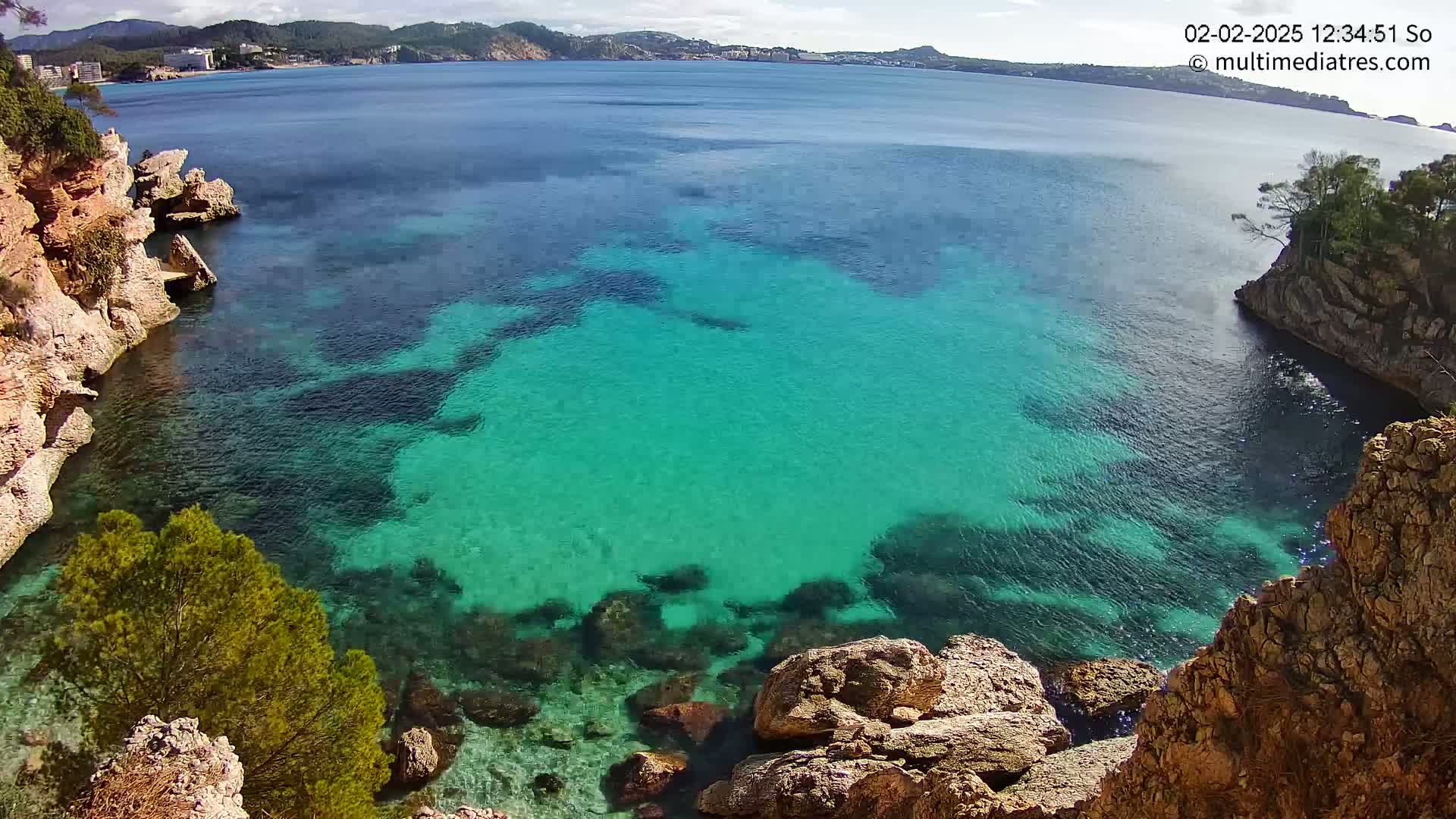 A cove of clear, teal water is nestled between rocky cliffs on a sunny day, with a distant shoreline visible in the background.
