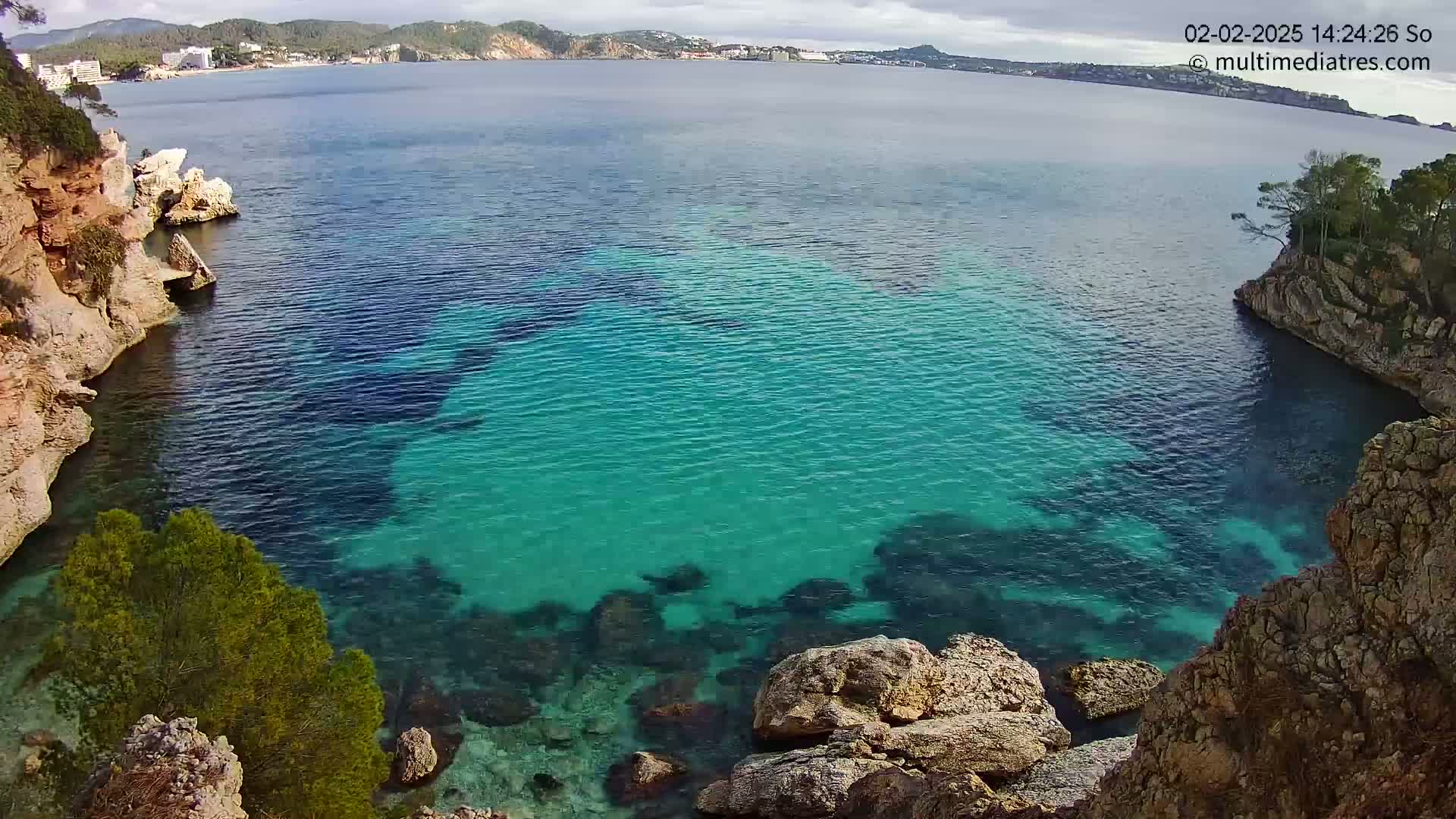 A cove of clear, teal water is nestled between rocky, tan-colored cliffs, with a distant coastline visible under a partly cloudy sky.
