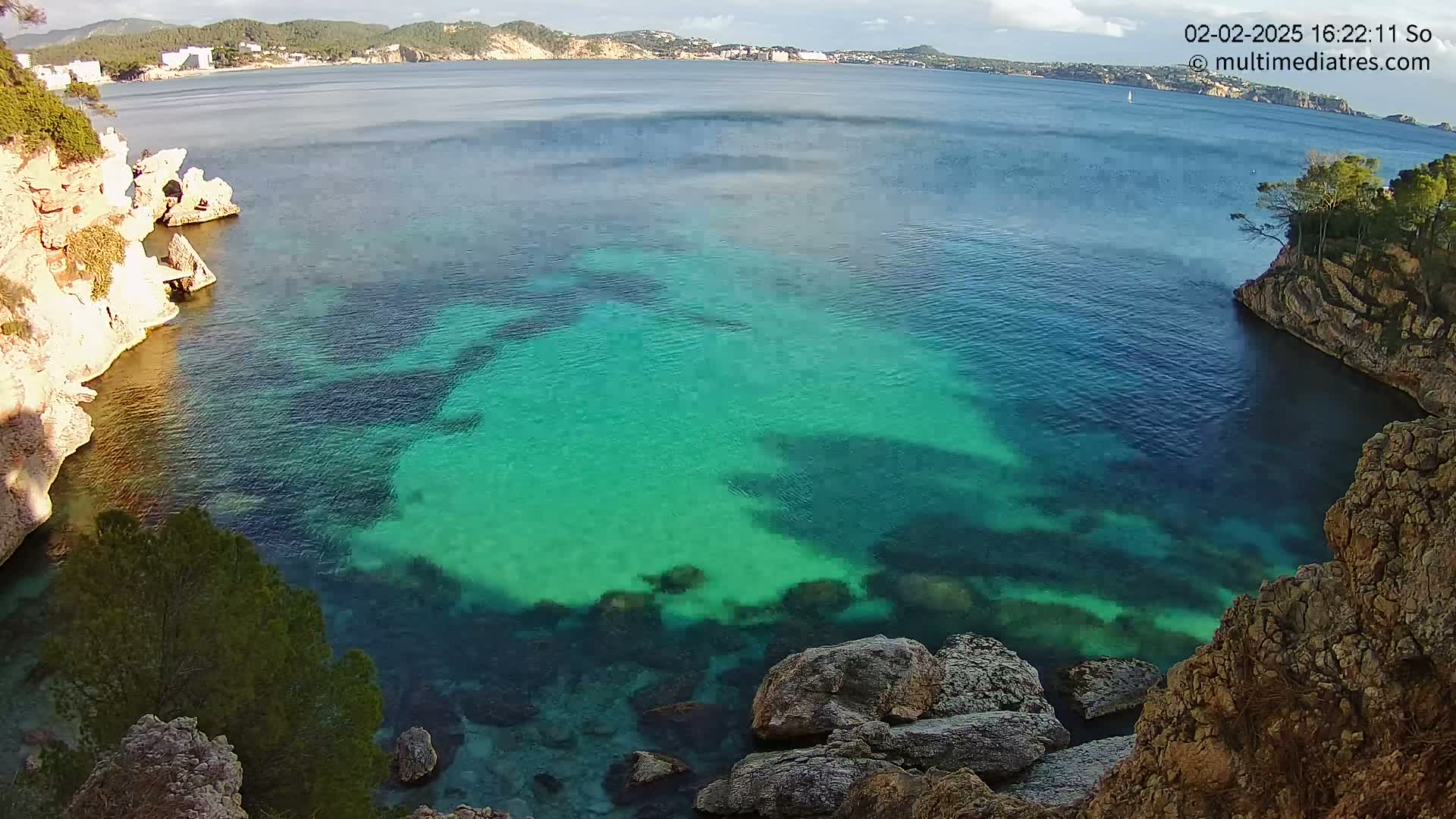 Clear, teal water fills a cove between rocky cliffs, with a calm sea and distant coastline visible in the background under a partly cloudy sky.