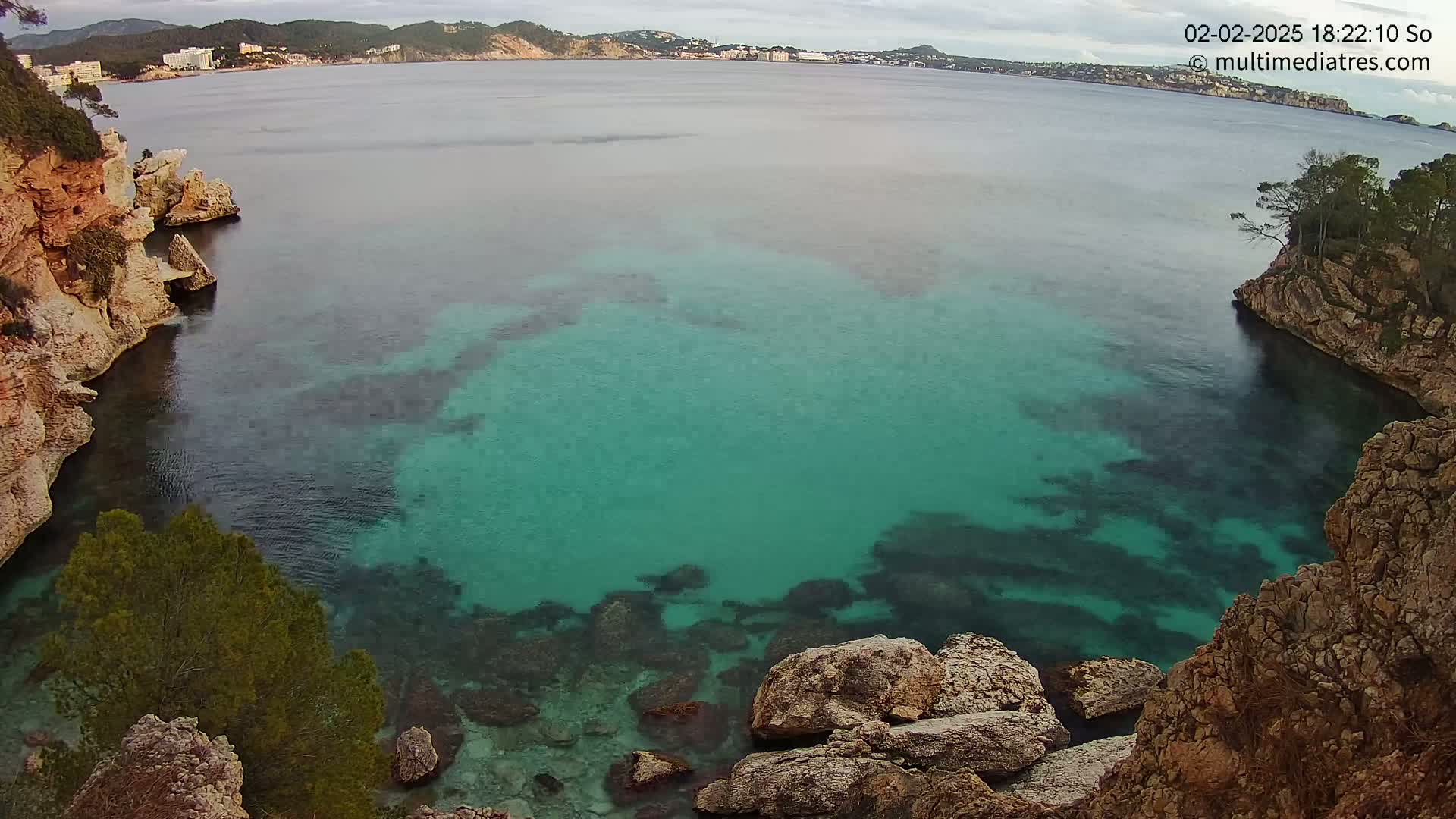 A cove of clear, teal water is nestled between rocky cliffs, with a distant town visible on a cloudy day.