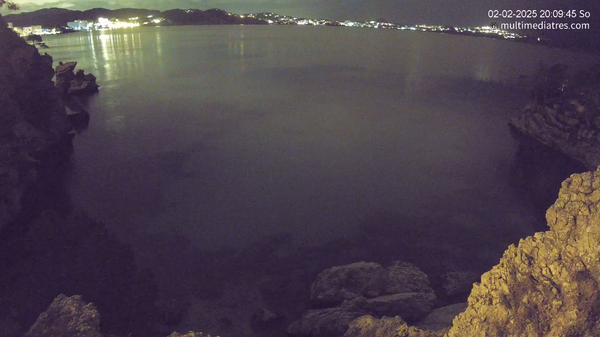 A nighttime view of a calm body of water reflecting the lights of a town on the opposite shore, with rocky cliffs on either side of the foreground.