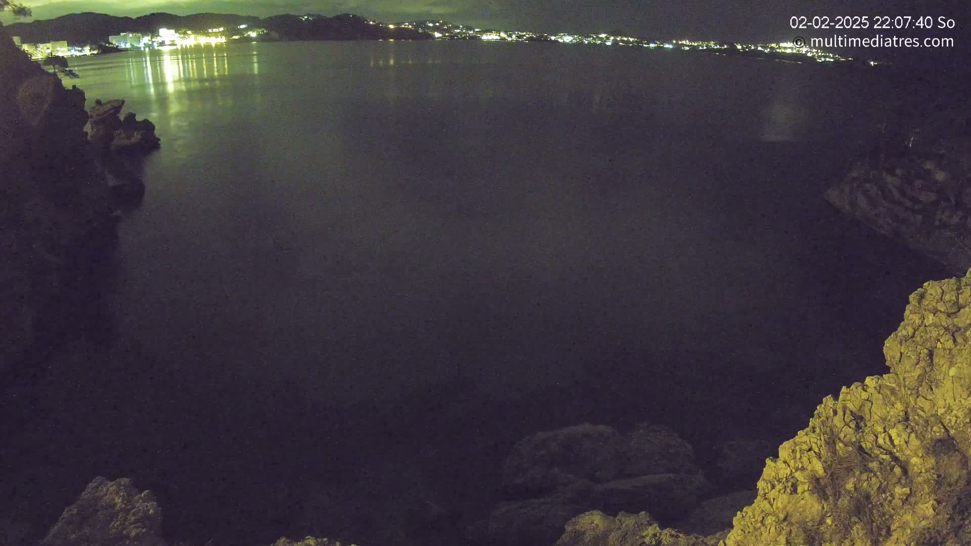 A nighttime view of dark water reflecting lights from a distant town, seen from a rocky cliff.