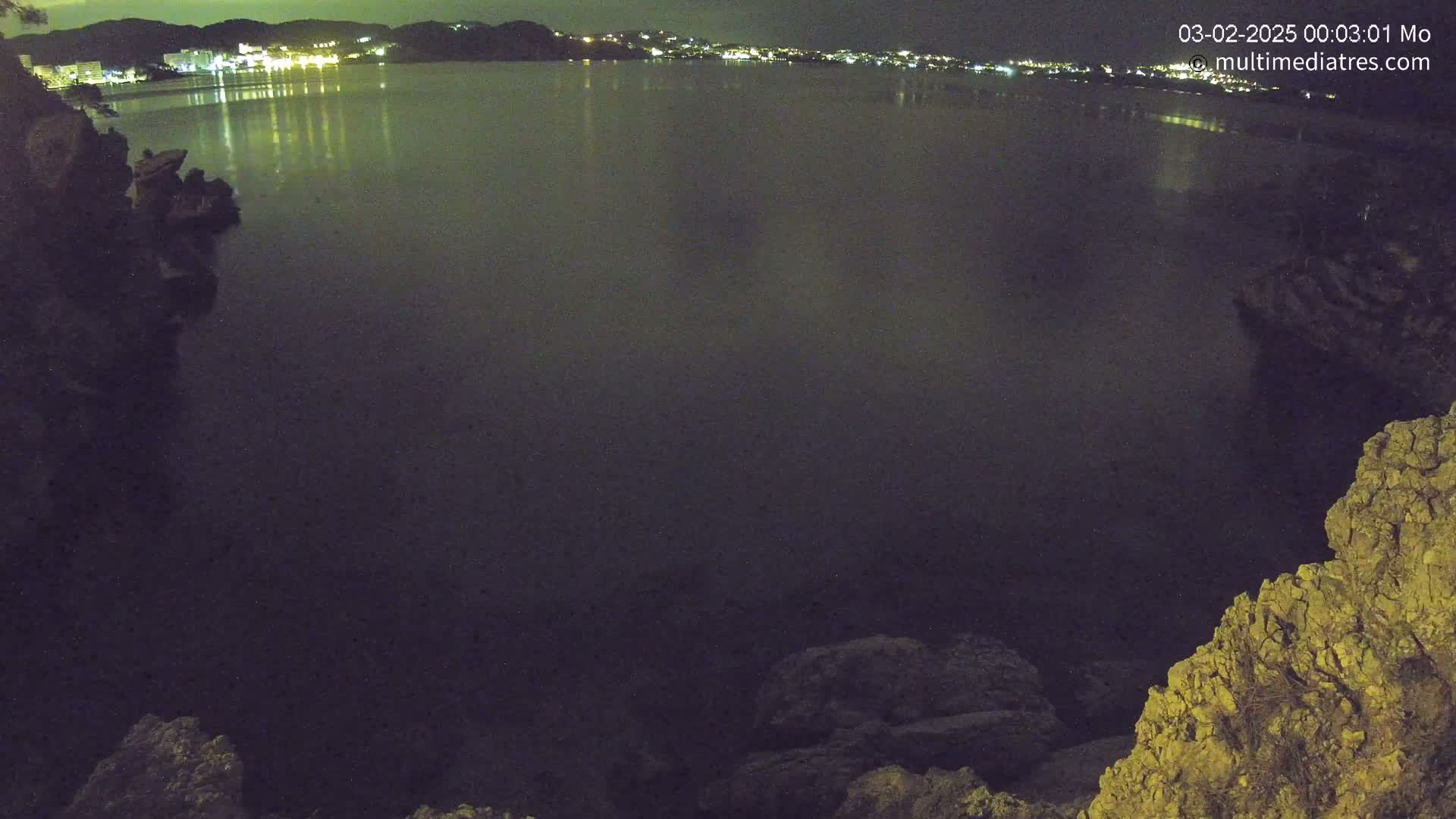 A dark body of water at night, reflecting the lights of a town in the distance, is viewed from a rocky overlook.