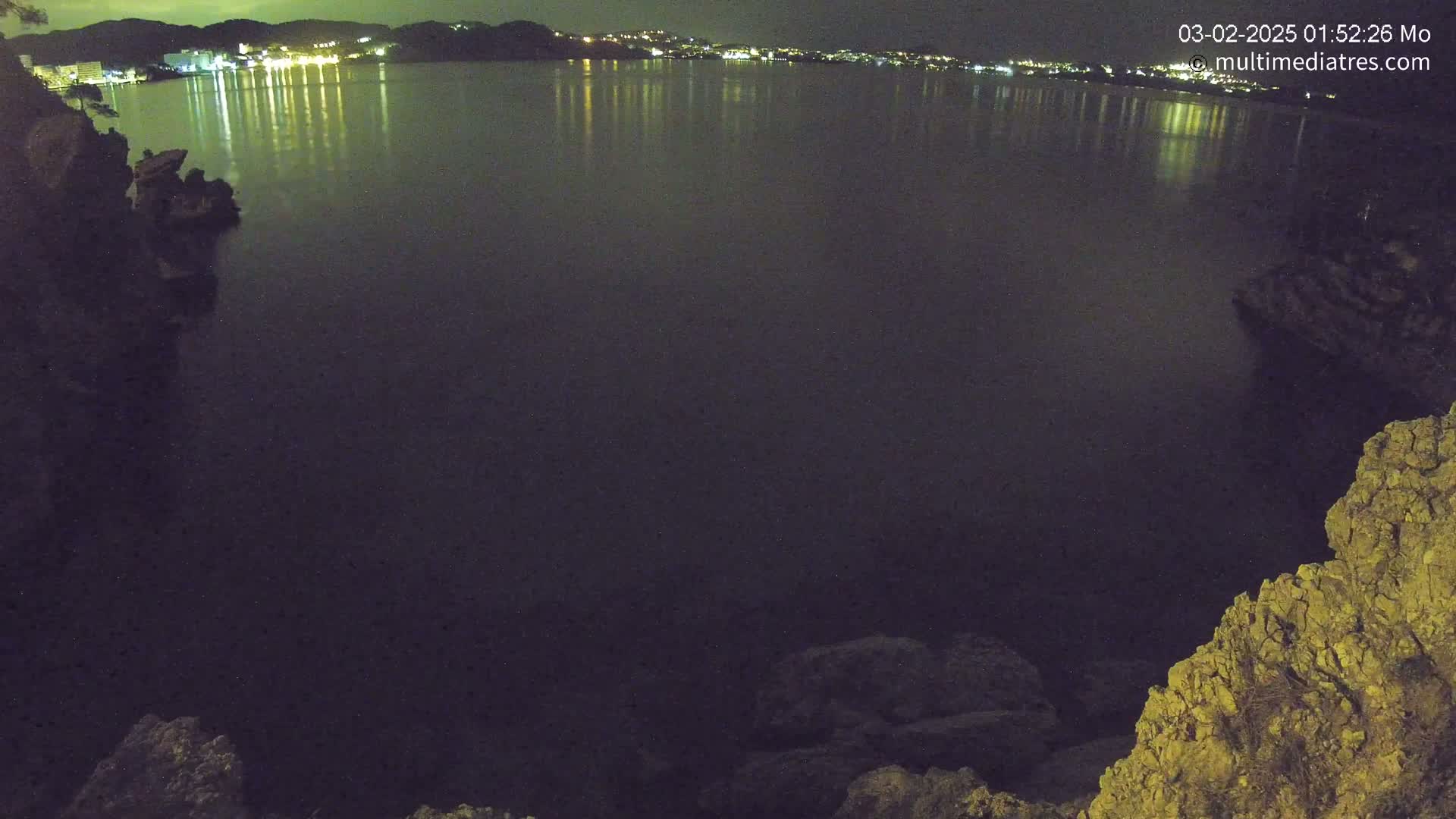 A dark, calm body of water reflects the lights of a town across its surface at night, viewed from a rocky vantage point.