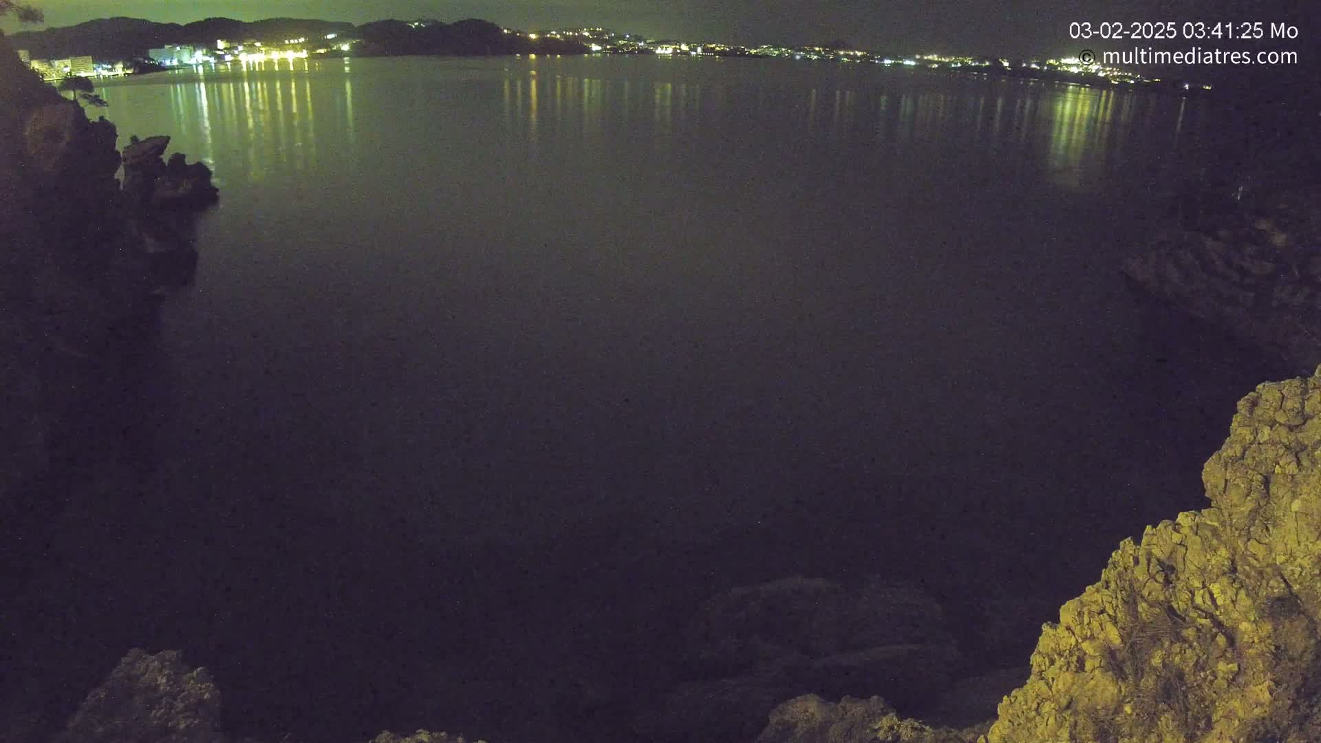 A nighttime view of a dark body of water reflecting the lights of a distant town, seen from a rocky vantage point.