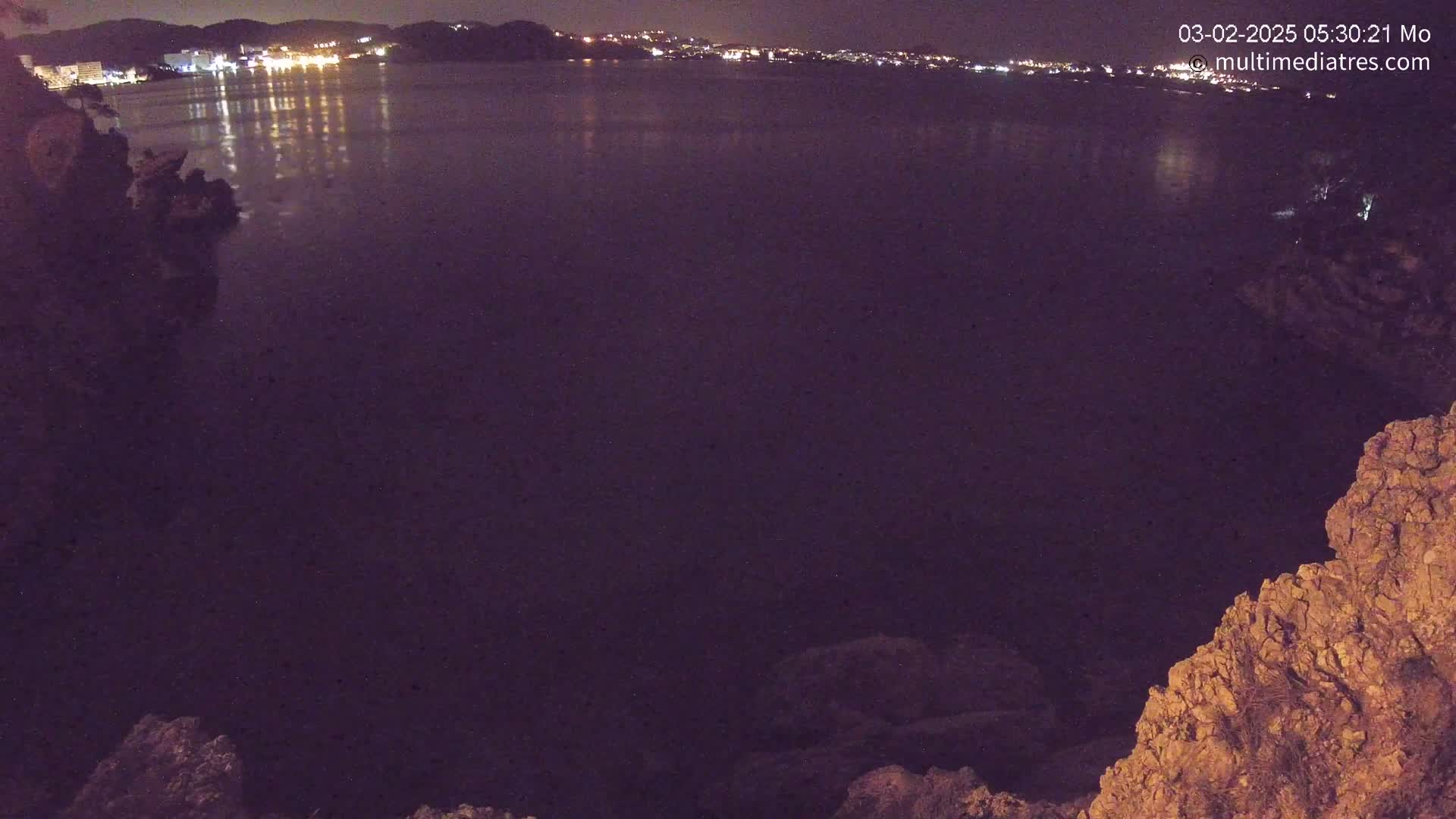 A nighttime view of a calm body of water reflecting the lights of a town across the water, seen from a rocky vantage point.