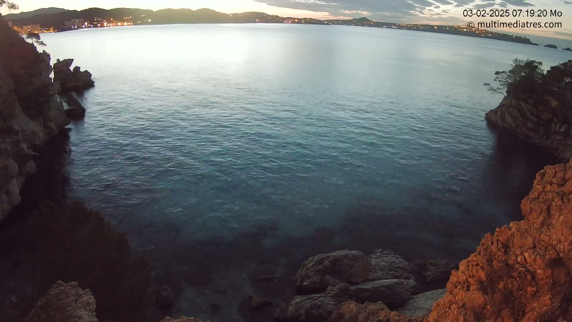 A calm, dark-blue sea is nestled between rocky, reddish-brown cliffs at dusk, with a distant town illuminated by lights visible across the water.