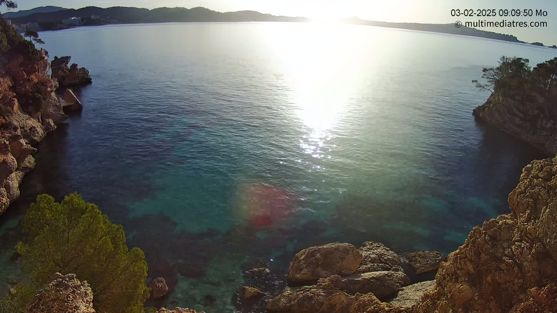 A cove of clear, blue-green water is nestled between rocky cliffs on a sunny morning.