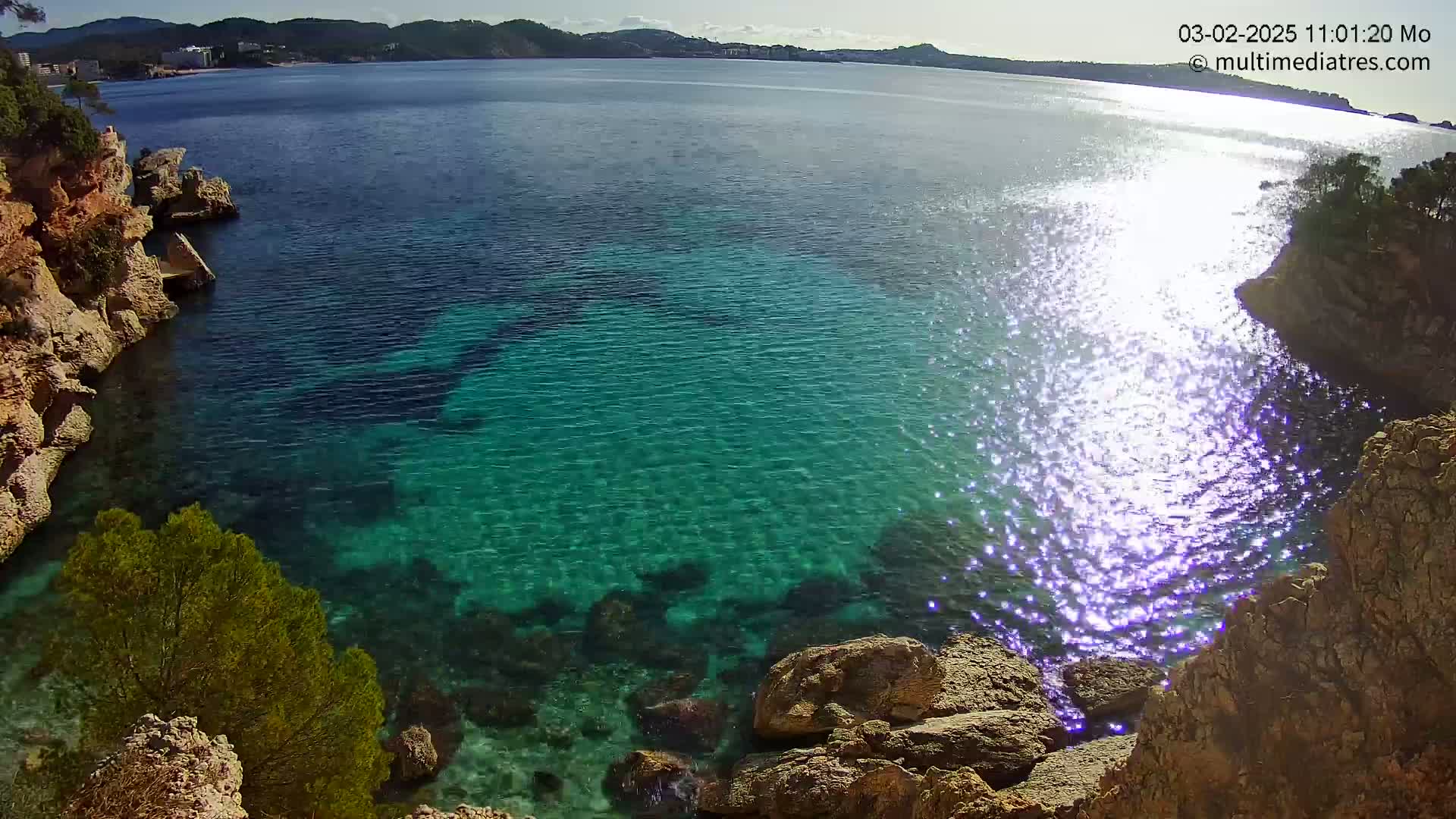 Clear, sparkling turquoise water laps against rocky shores under a bright, sunny sky.