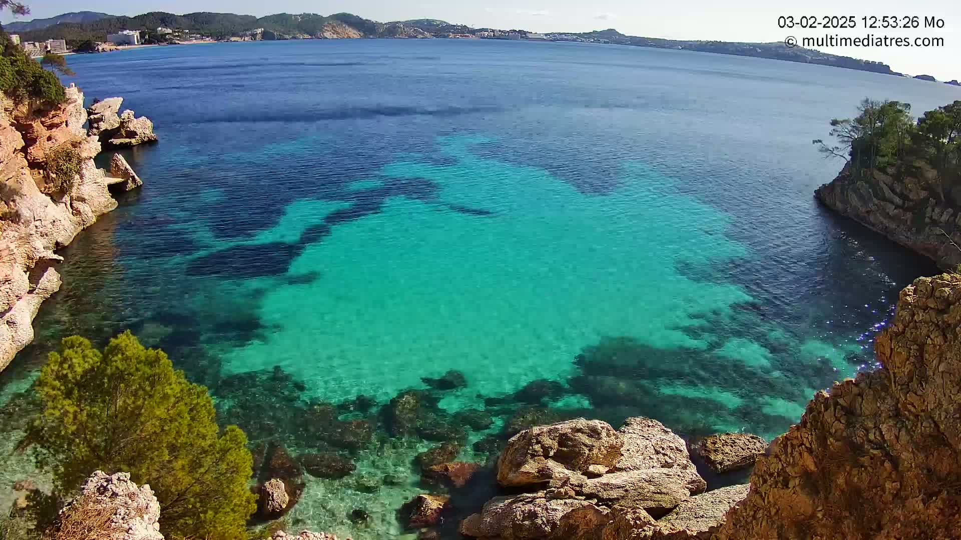 Clear, turquoise water fills a cove surrounded by rocky, tan cliffs and green vegetation under a sunny sky.