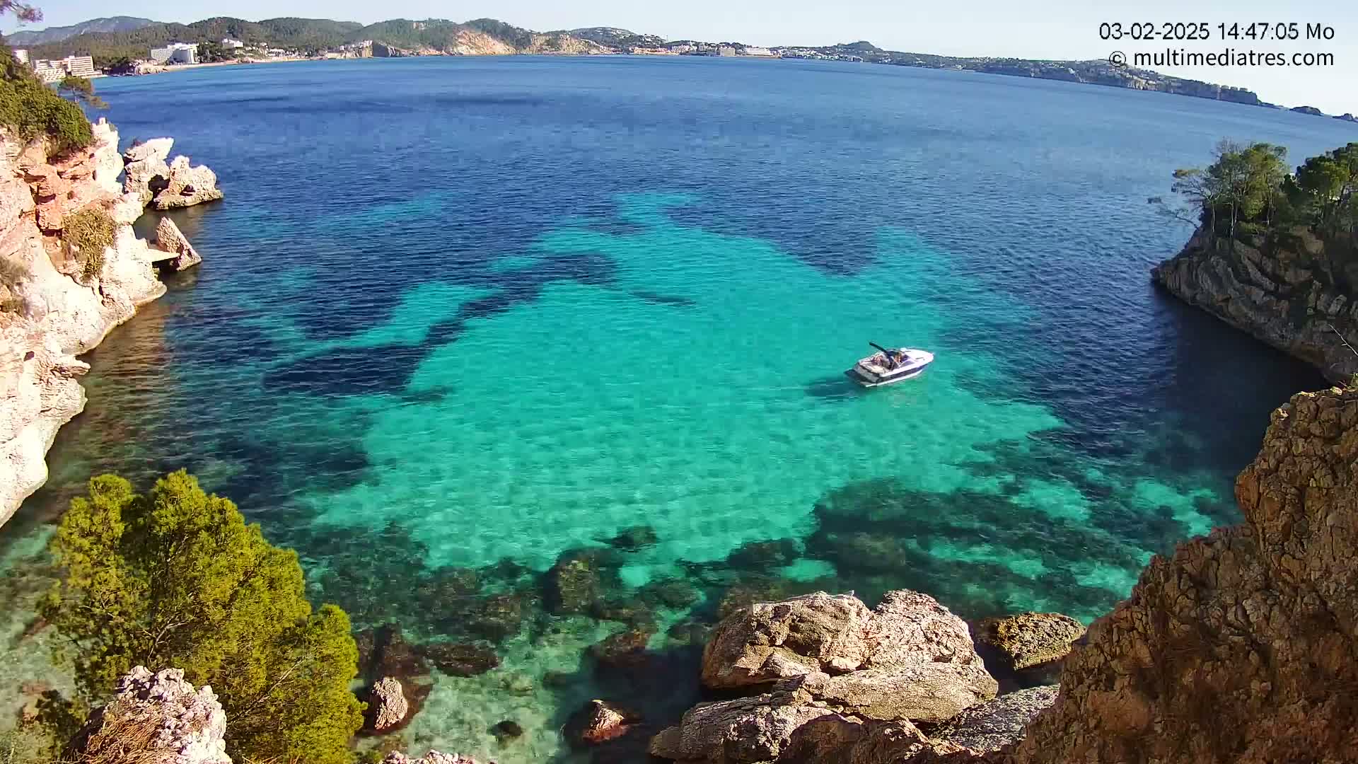 A small boat sits in a calm, clear turquoise cove surrounded by rocky cliffs and vegetation on a sunny day.